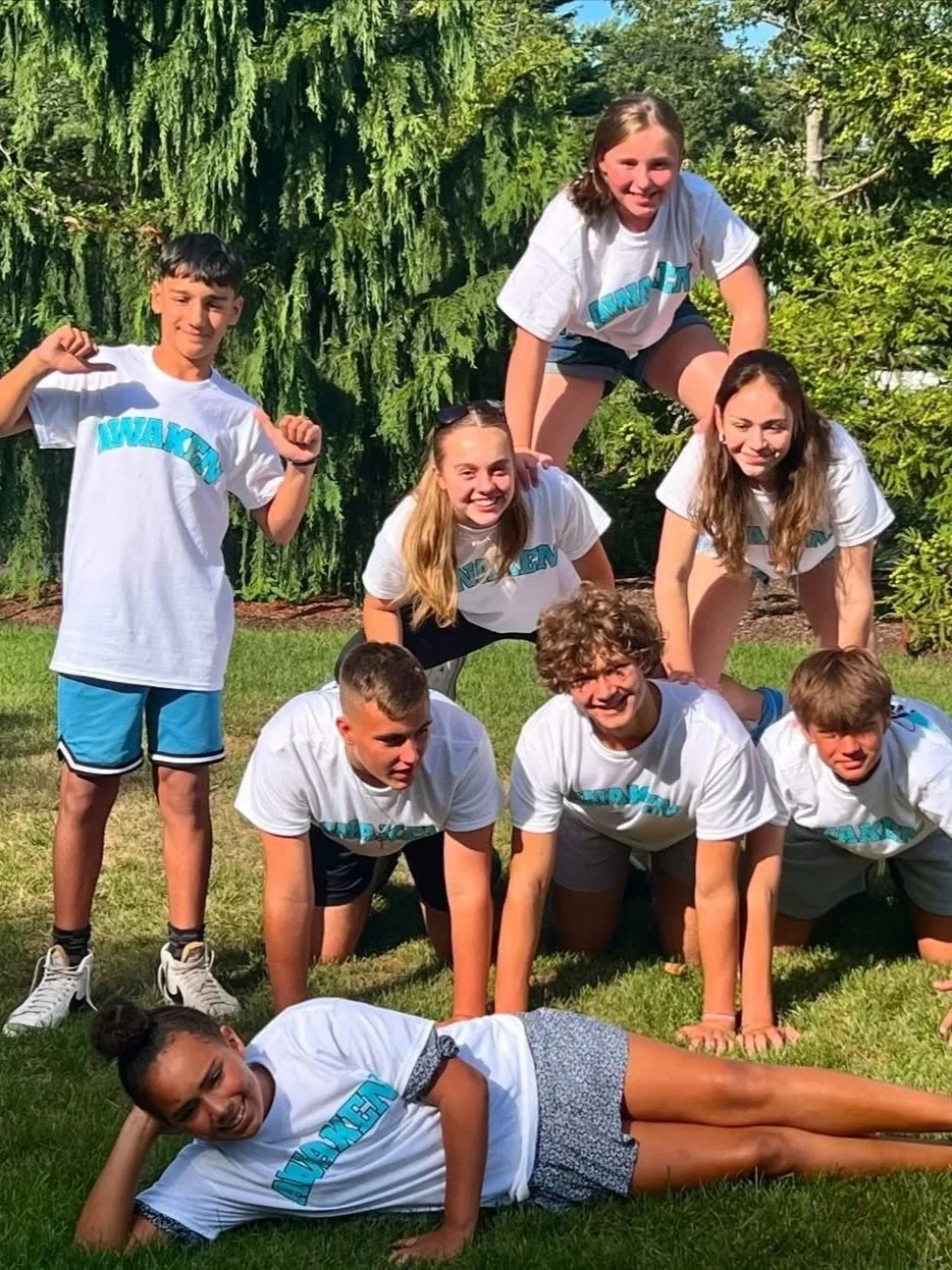 A group of nine children forming a human pyramid outdoors on grass, with a girl at the top, three girls in the middle, four boys supporting at the bottom, and a girl lying on the grass in front, all smiling and wearing white T-shirts with blue text.