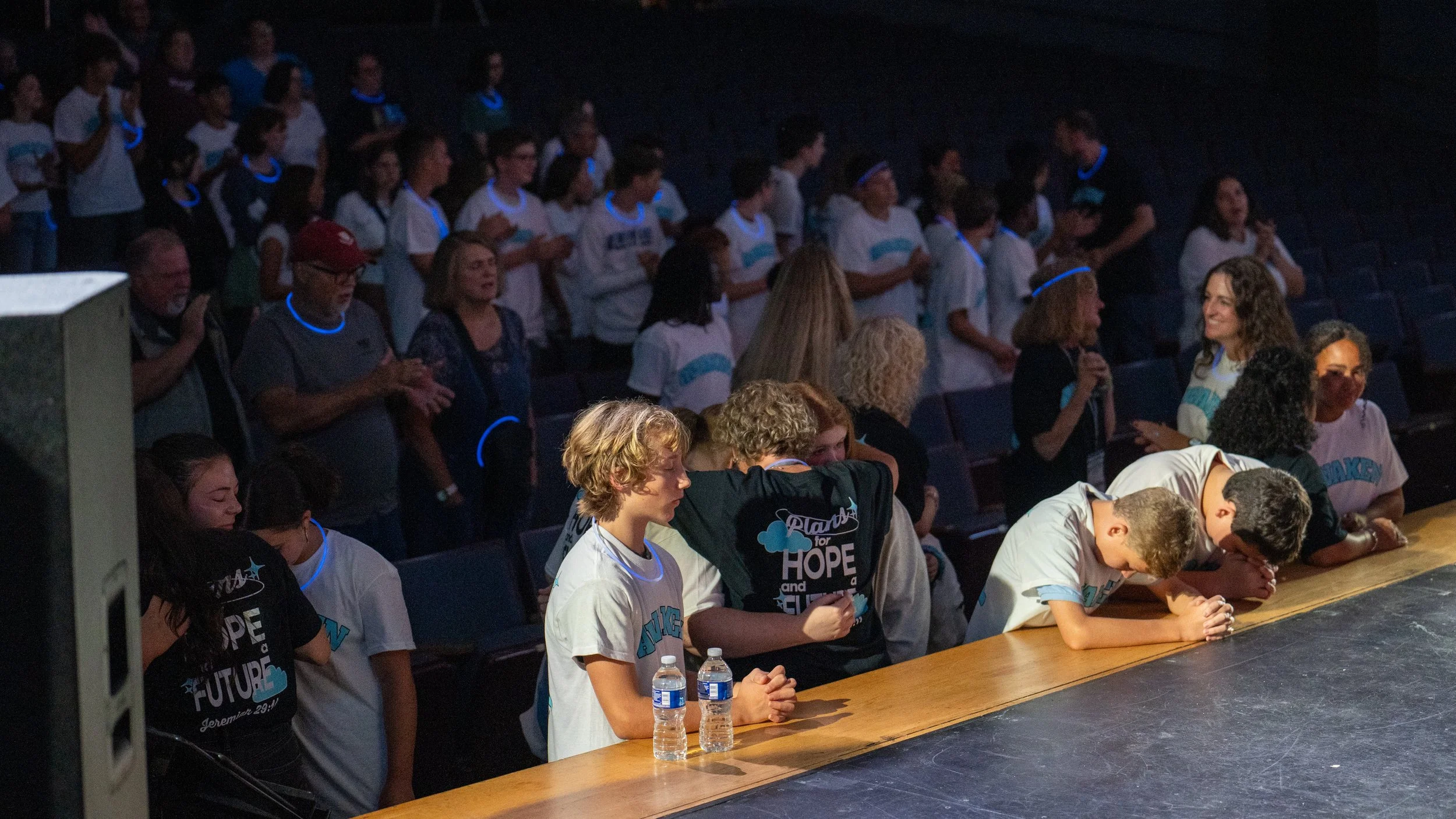 A group of young people and adults gathered in an auditorium, some sitting at a table with their hands clasped and heads bowed, while others in the background are standing and clapping, wearing event T-shirts and glow necklaces.