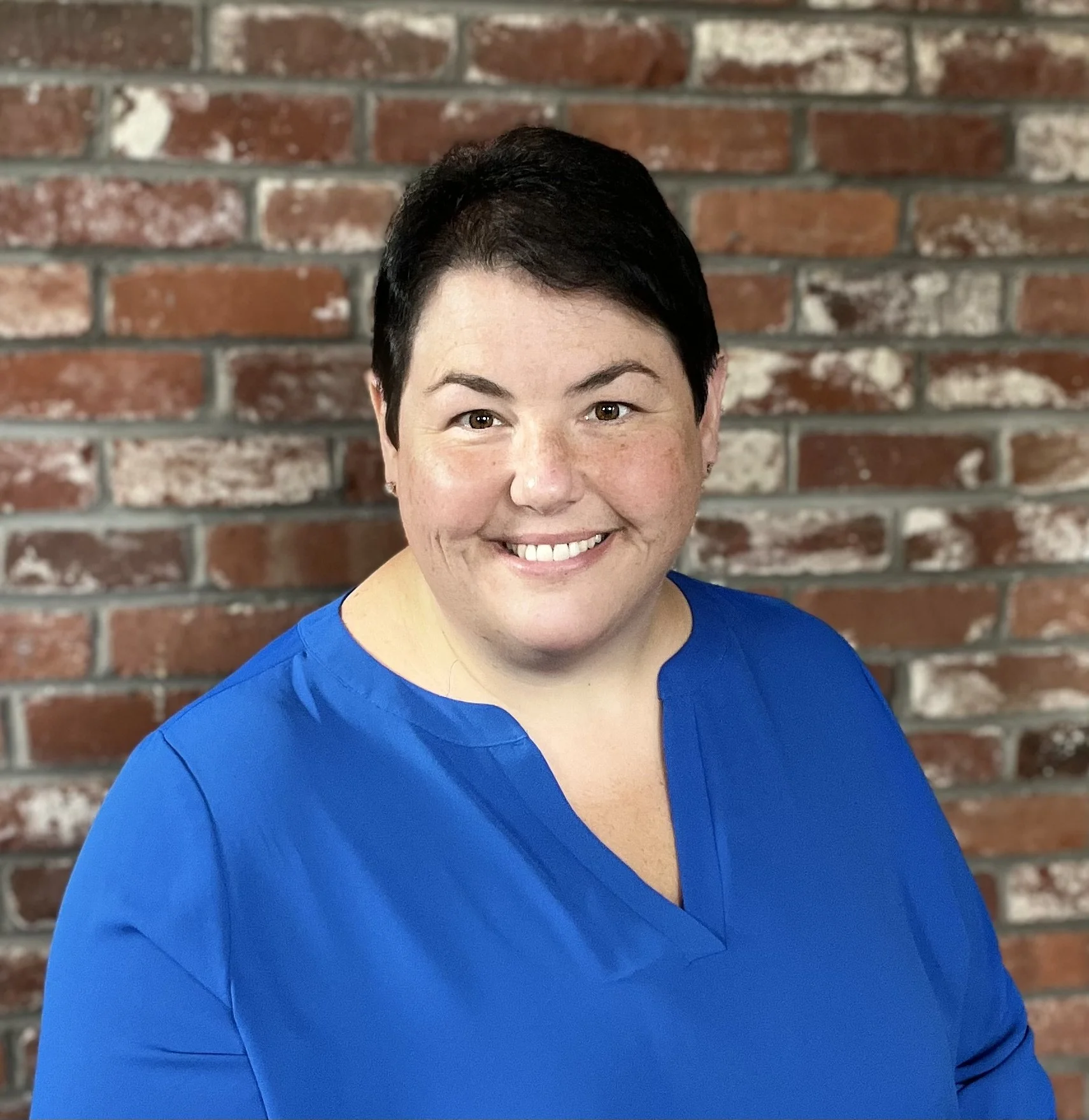 Smiling woman with short dark hair wearing a bright blue top, standing in front of a red brick wall.