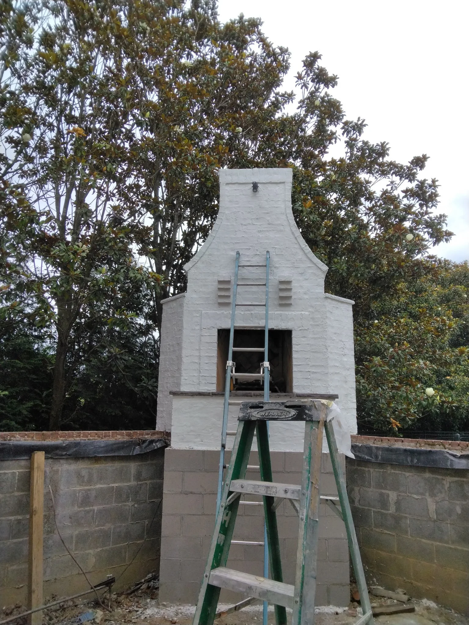 A white-painted brick outdoor fireplace or chimney structure in the process of construction or renovation, with a ladder leaning against it, set in a backyard with a tree in the background.