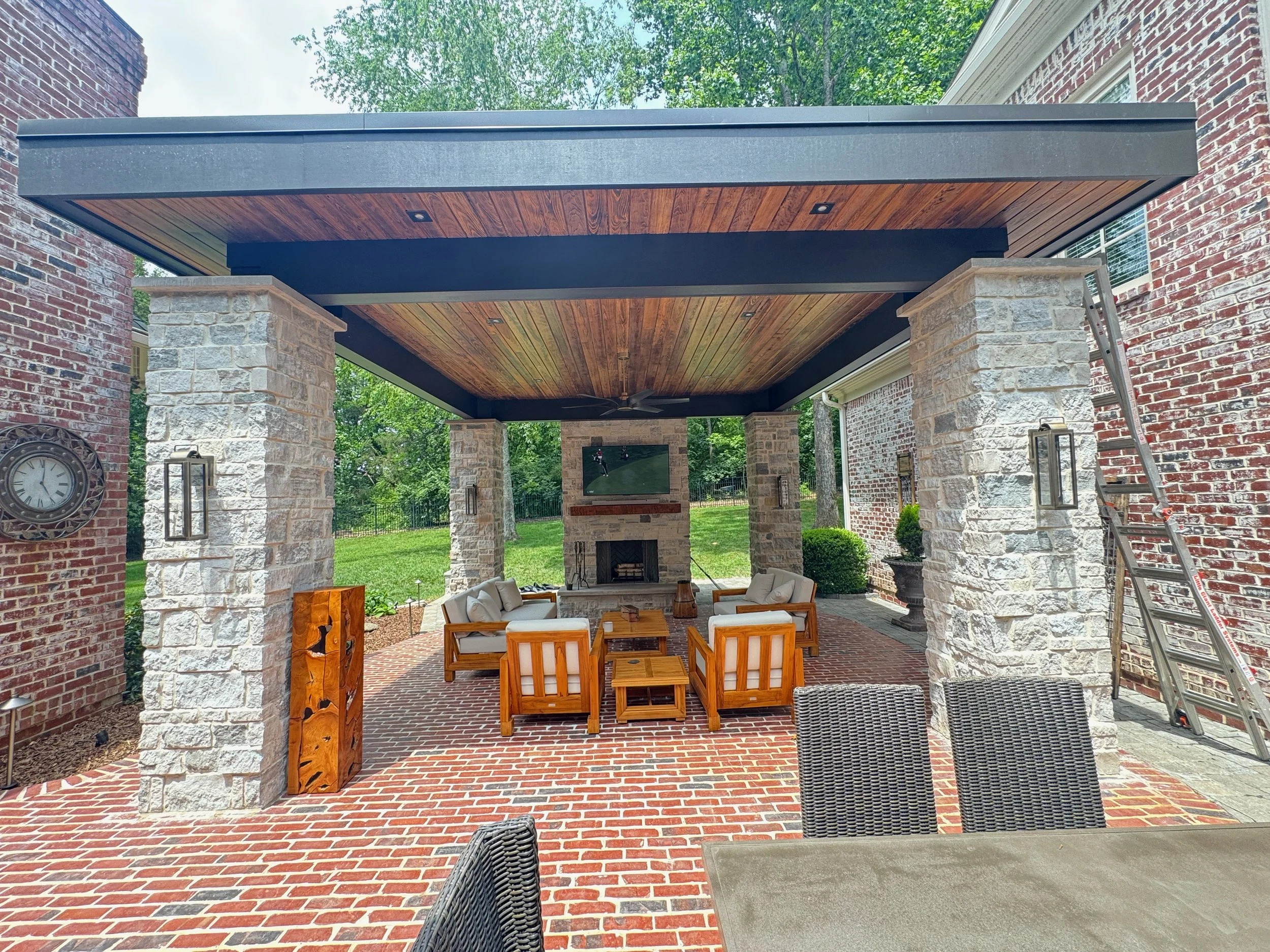 Covered patio area with brick flooring, stone columns, ceiling fan, and outdoor seating. A wall-mounted TV, clock, and a ladder are visible. The ceiling has wood paneling and recessed lighting. The background shows green trees and a grassy yard.