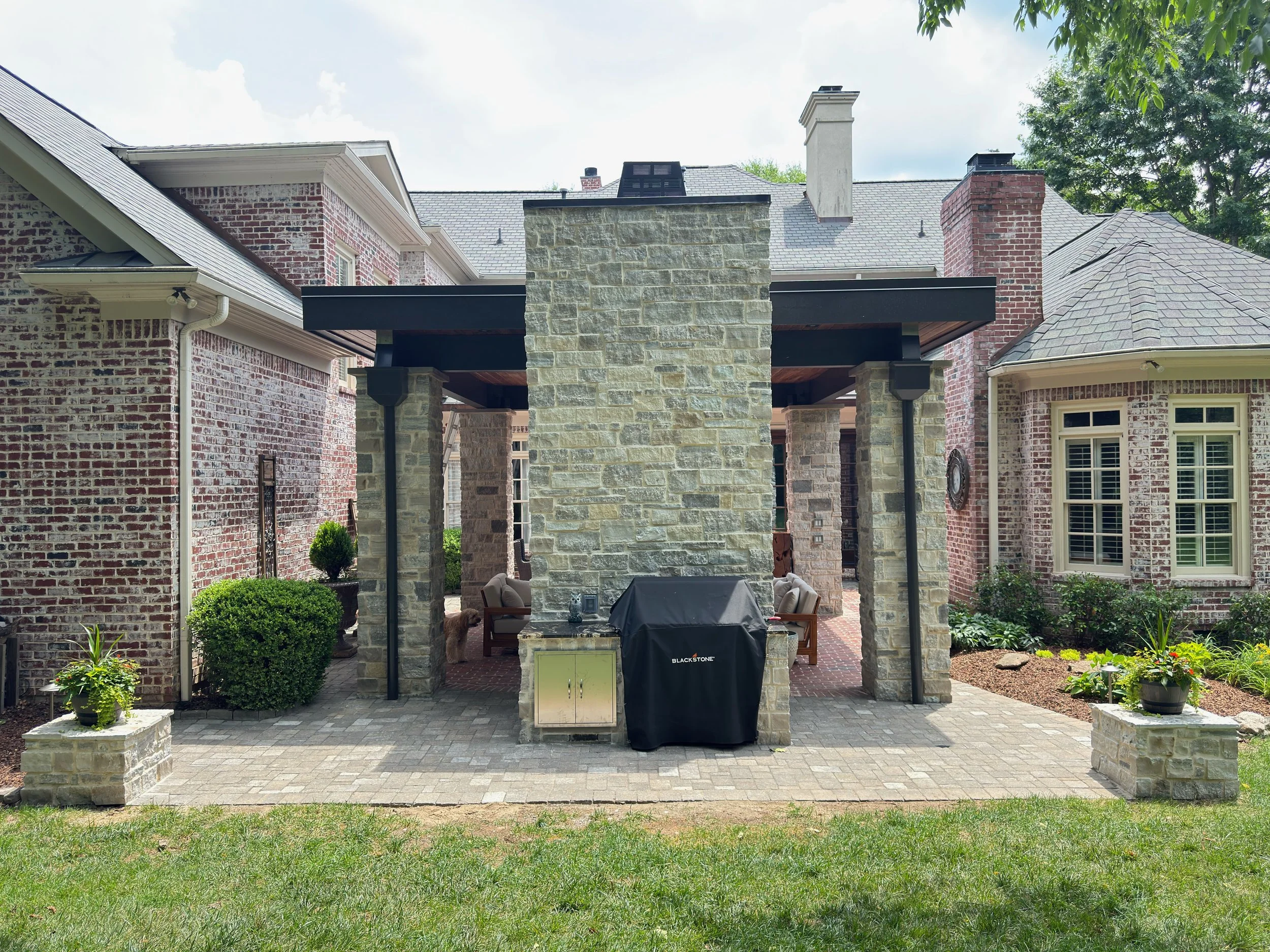 Backyard patio with brick and stone exterior walls, a chimney, a covered sitting area with patio furniture, and greenery in the garden.