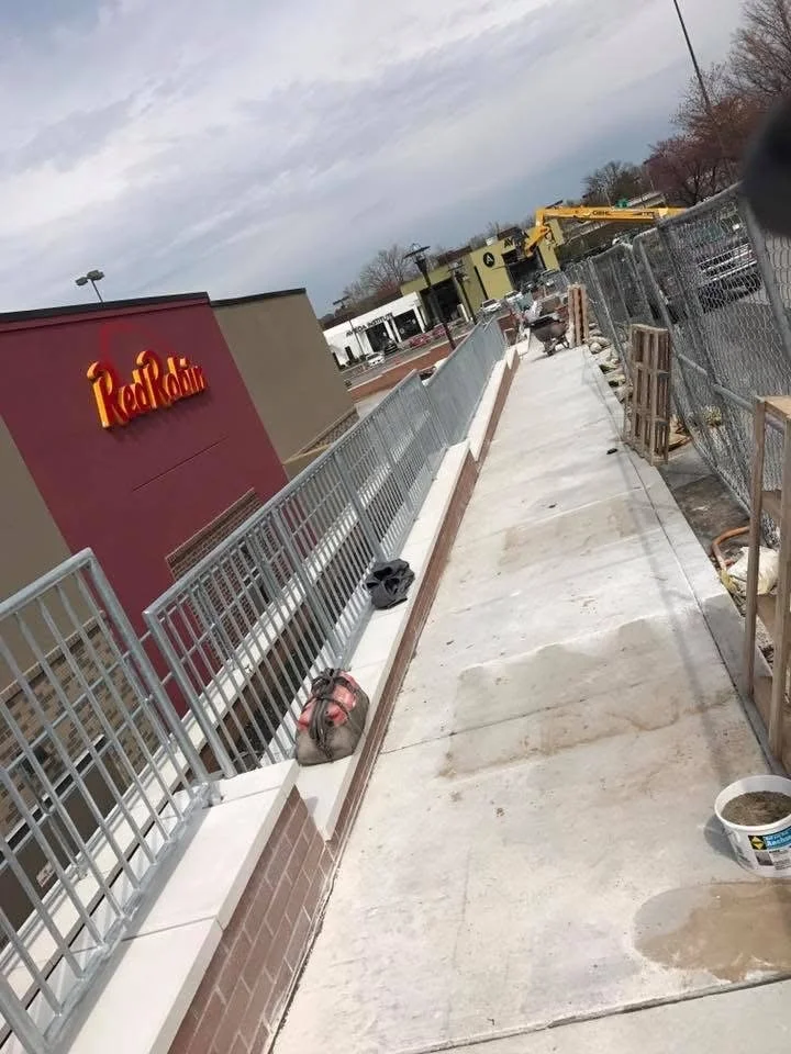 Sidewalk under construction outside a Red Robin restaurant, with construction materials and equipment along the sidewalk and a chain-link fence separating the sidewalk from the construction area. The sky is cloudy.