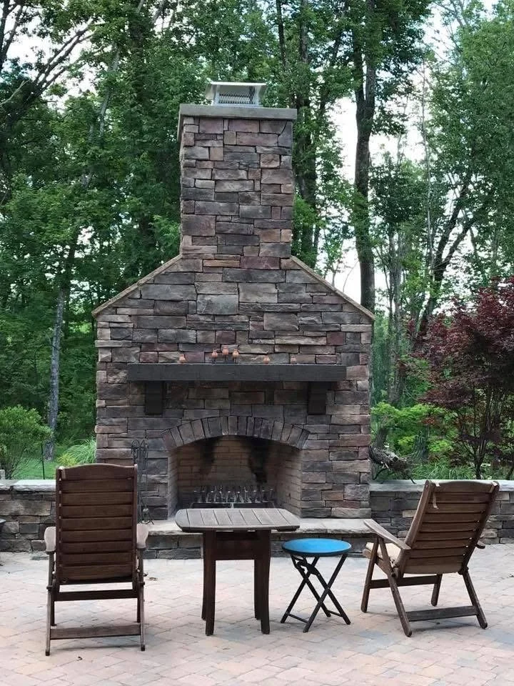 Outdoor stone fireplace with chimney surrounded by trees, two wooden chairs, a small wooden table, and a folding blue stool on a brick patio.
