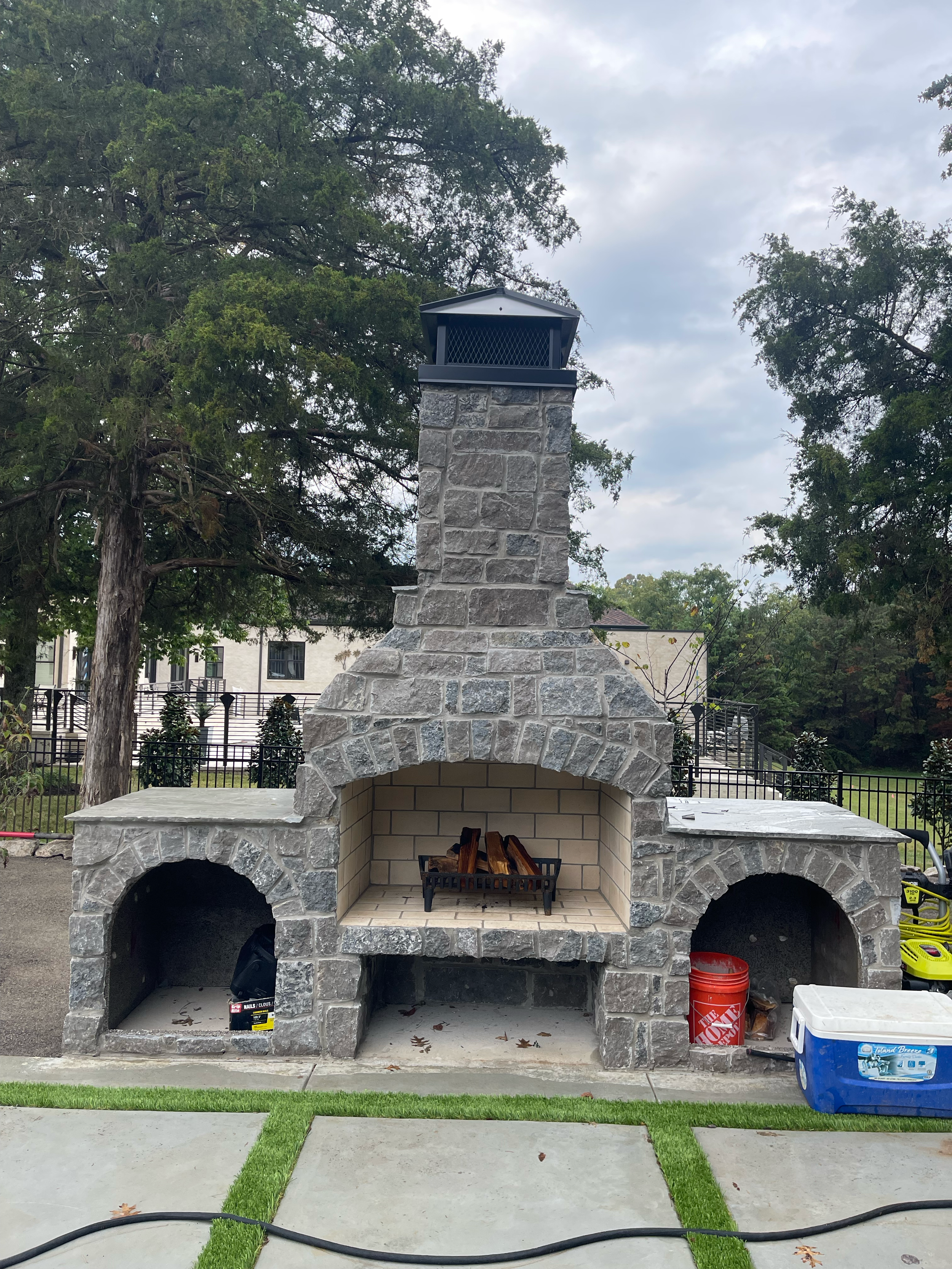 An outdoor stone fireplace with a chimney, set on a patio with grass and concrete. The fireplace has a wood storage area underneath and a metal shelf with firewood inside. To the right, there is a red bucket and a blue cooler. The background features trees and a black fence.
