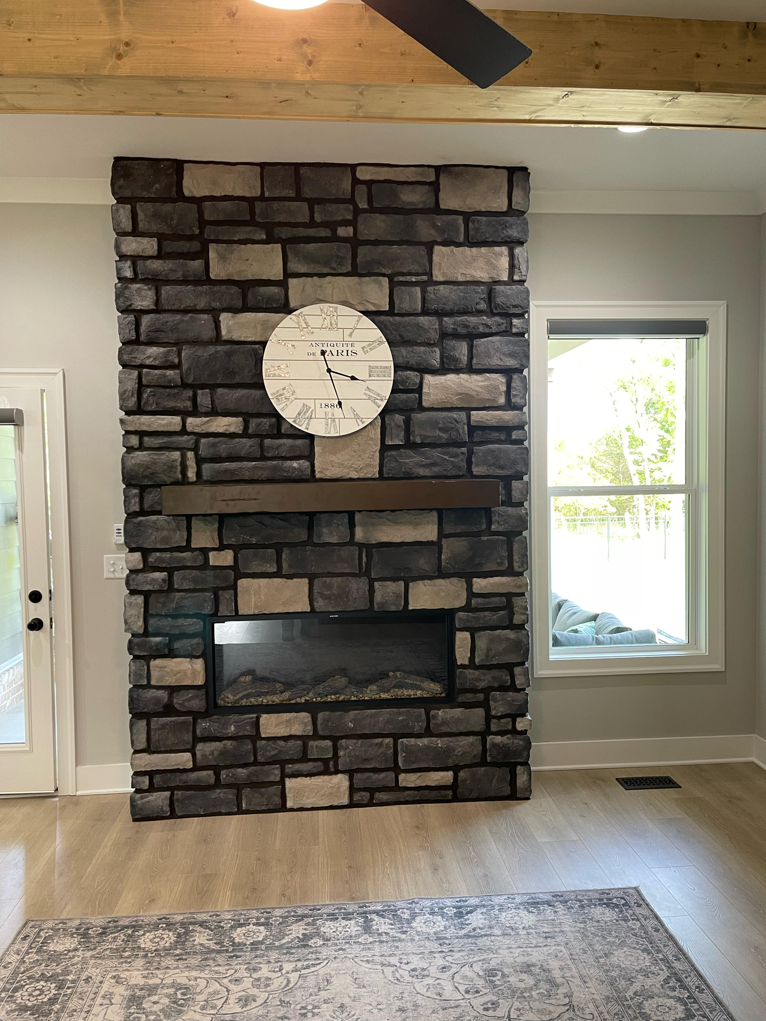 Interior view of a living room with a black and beige stone fireplace, a round wall clock, a window to the right, a floor vent, and a section of an area rug on light wood flooring.