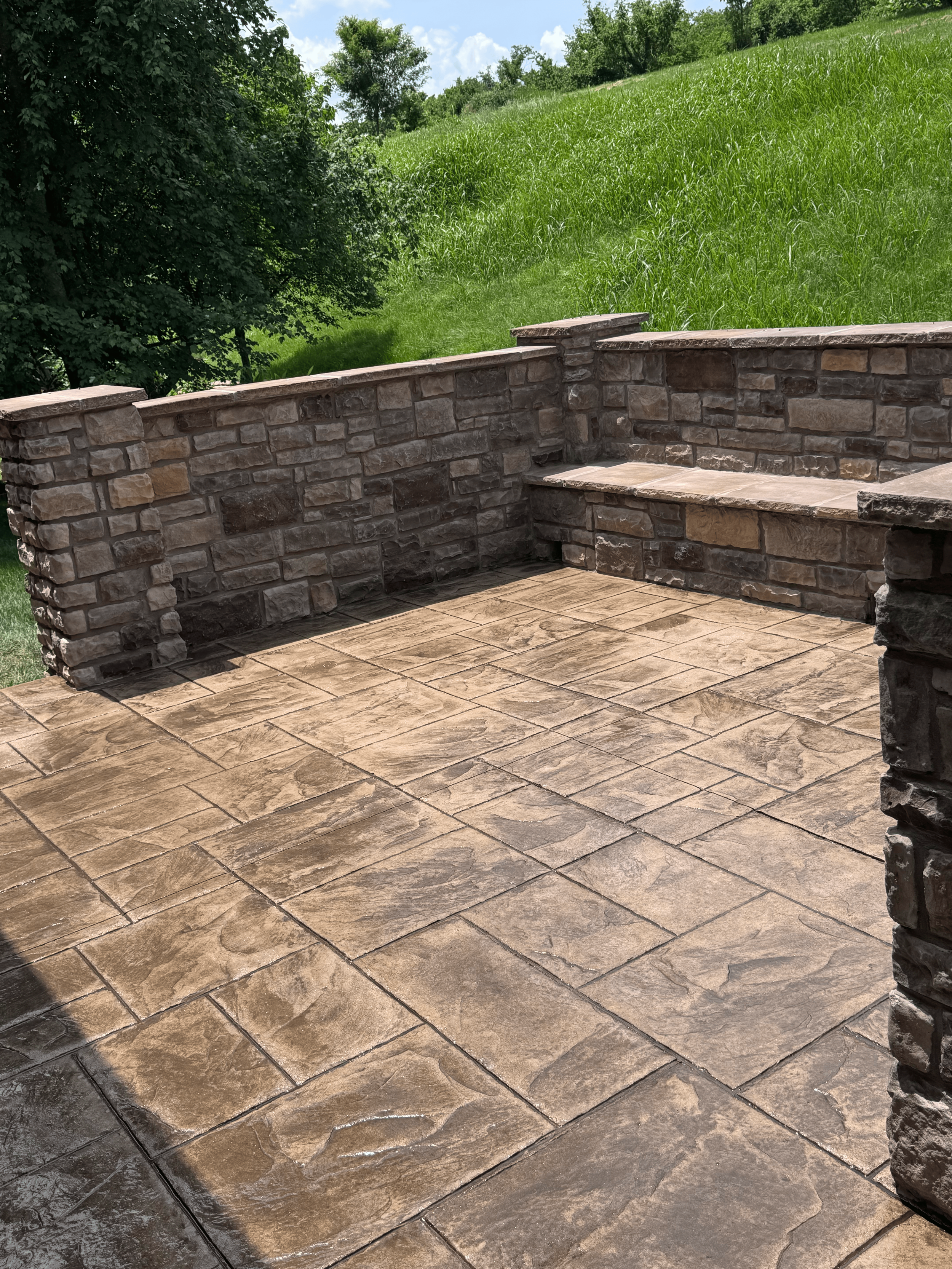 A small outdoor patio with brown stamped concrete flooring and a low stone wall, near a grassy hill with trees and a partly cloudy sky in the background.