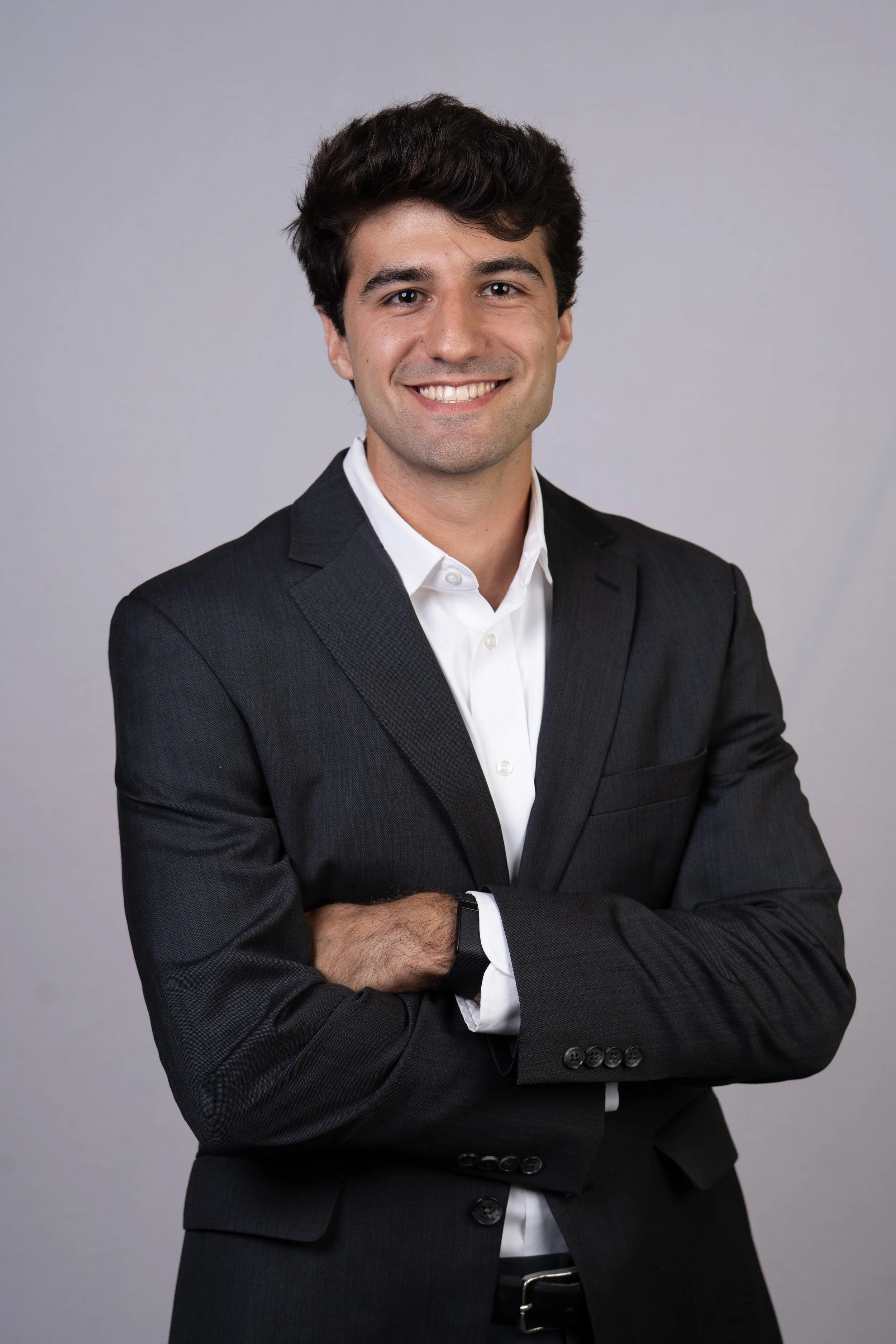Portrait of a young man in a black suit with arms crossed, smiling, against a plain gray background.