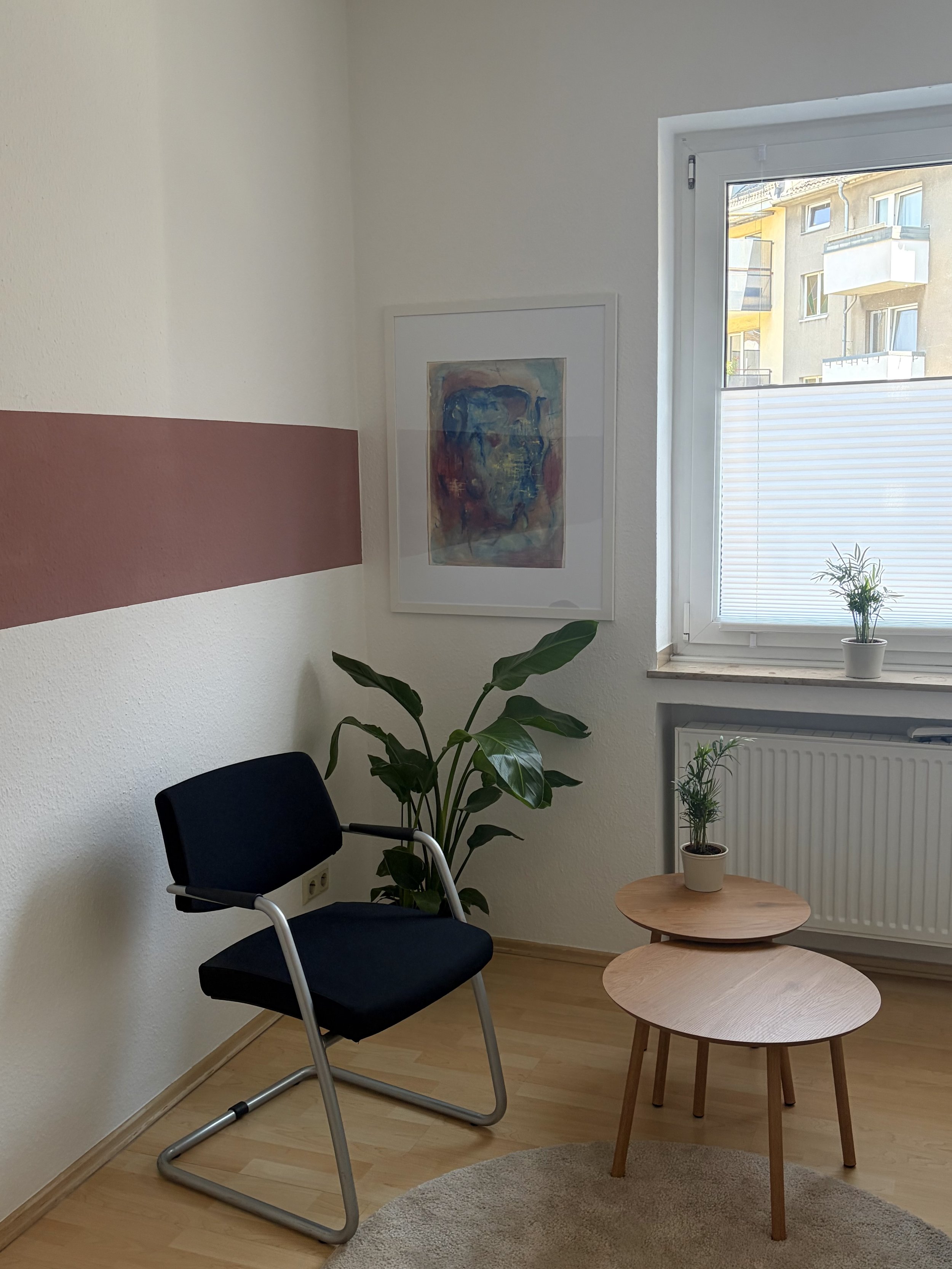 Ein Praxisraum mit weißen Wänden, einem Fenster, ein dunkel Stühle, zwei kleinen runden Holztischen, einer Zimmerpflanze, einem Bilderrahmen,, einem Teppich