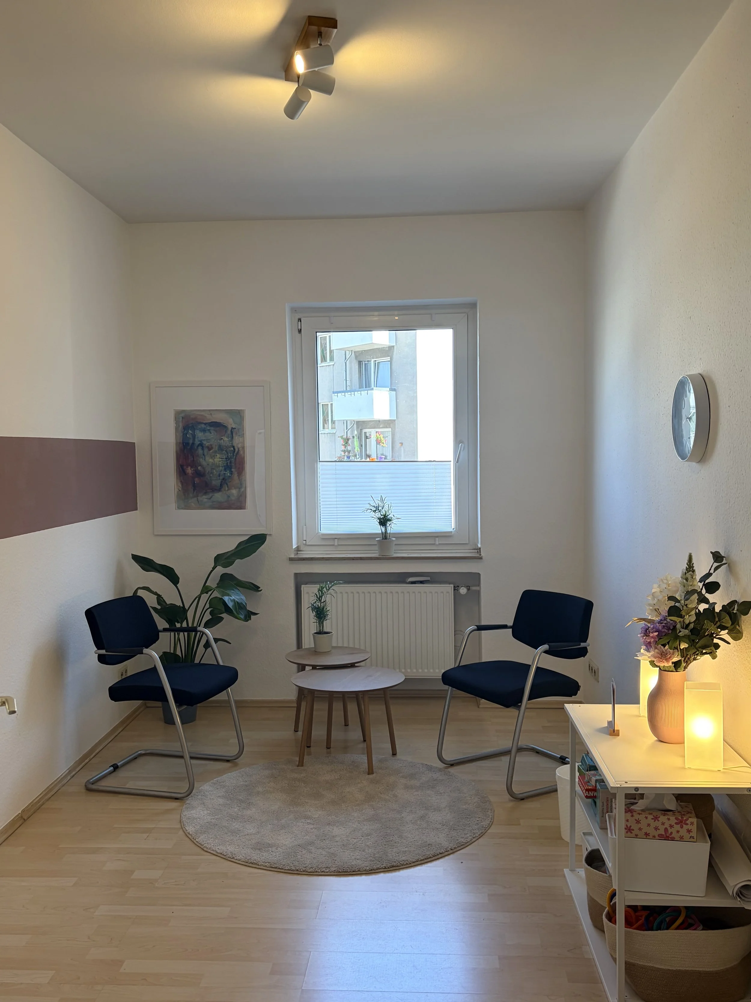 Ein Praxisraum mit weißen Wänden, einem Fenster, zwei dunkle Stühle, zwei kleinen runden Holztischen, einer Zimmerpflanze, einem Bilderrahmen, einer weißen Kommode mit Dekoration, einem Wanduhren, einem Teppich und einer Deckenleuchte