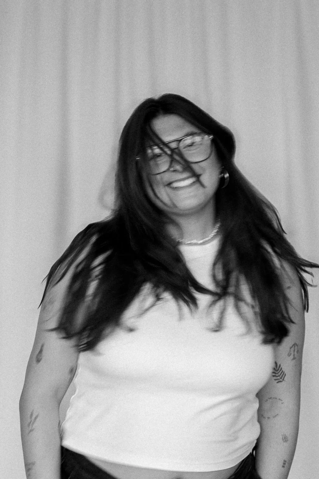 Junge Frau mit schwarzen Haaren, Brille, Tattoos am Arm, lächelnd, schwarze und weiße Fotografie, lässige Kleidung, Fröhlichkeit, Haare im Wind