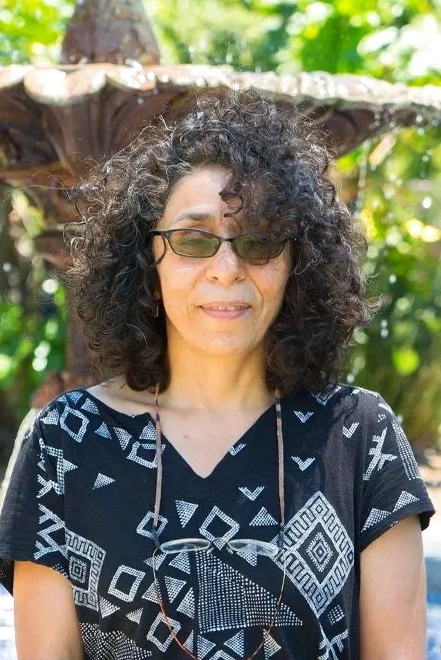 Mujer con cabello rizado y peinado voluminoso, usando gafas de sol oscuras y camiseta negra con patrones blancos, frente a fuente en un entorno exterior verde y soleado.