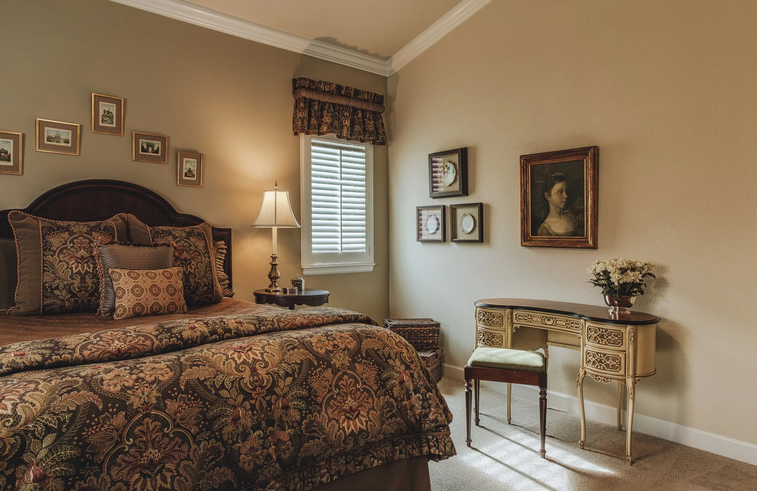 Interior design photo of a bed featuring a floral patterned bedspread and pillows, a nightstand with a lamp, a small window with blinds, framed artwork on the walls, and a vintage-style console table with a flower vase, taken in Longmont, Colorado.