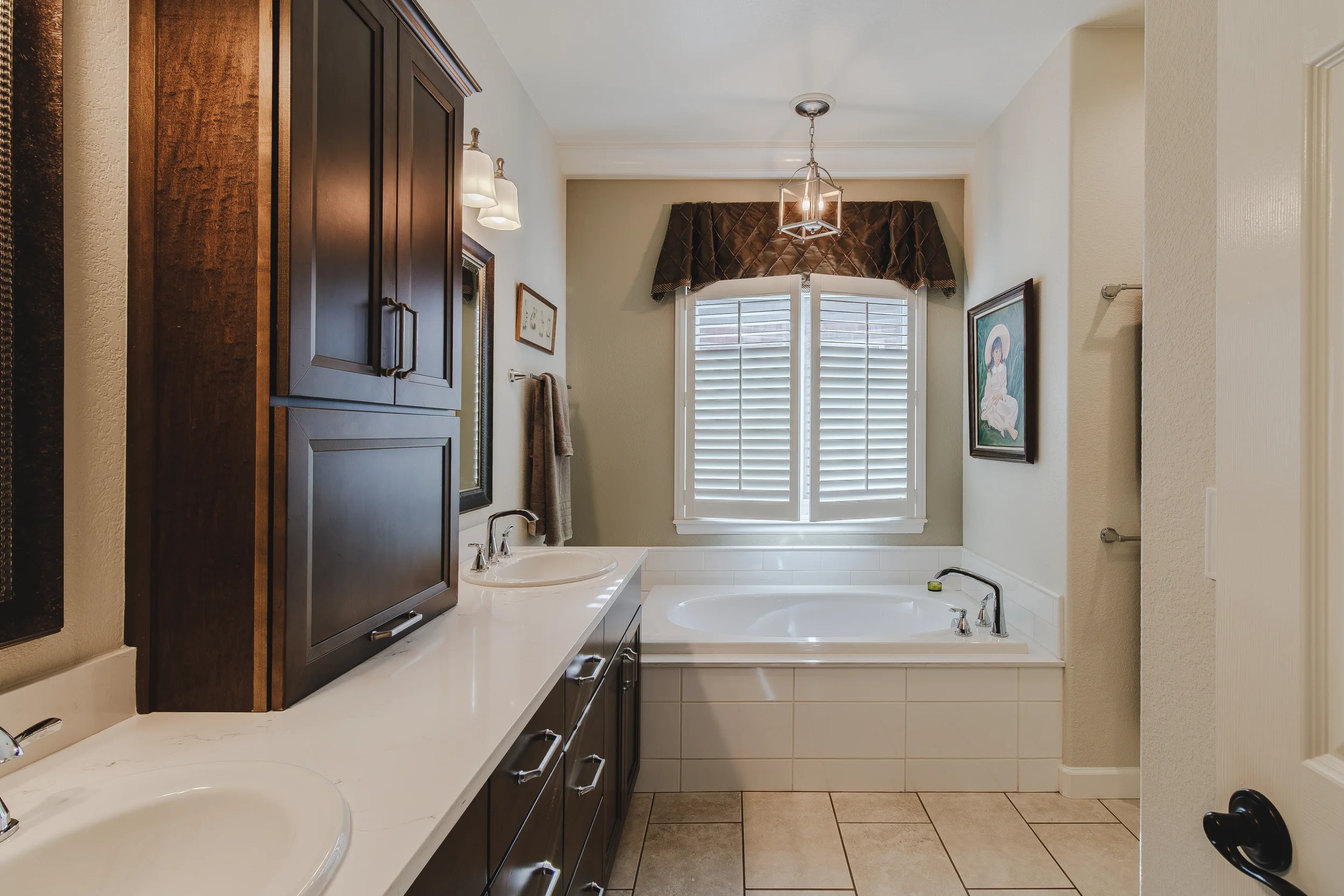Architectural photo of a bathroom with a double sink vanity, a bathtub by a window with shutters, framed art on the walls, and a ceiling light fixture, taken in Longmont, Colorado by Context Images. 