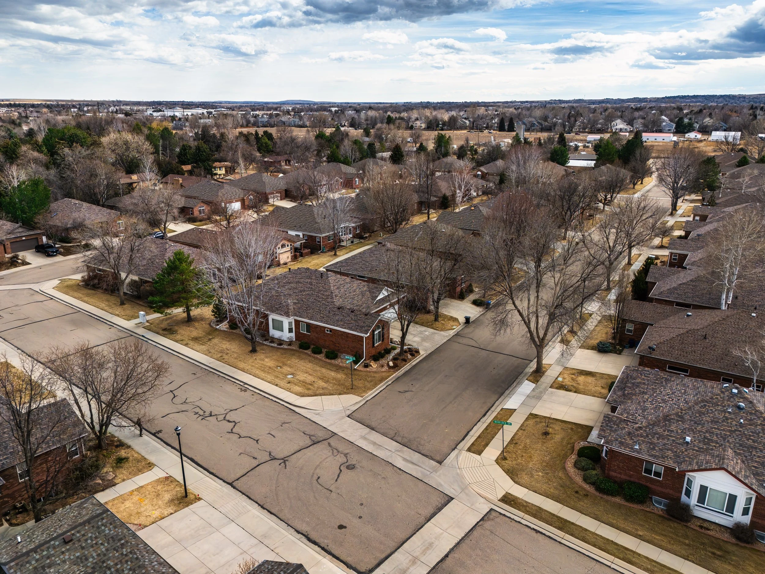 Aerial drone view of a suburban neighborhood with single-family homes, leafless trees, and streets with some cracks, under a cloudy sky. Taken in Longmont, Colorado by Context Images. 