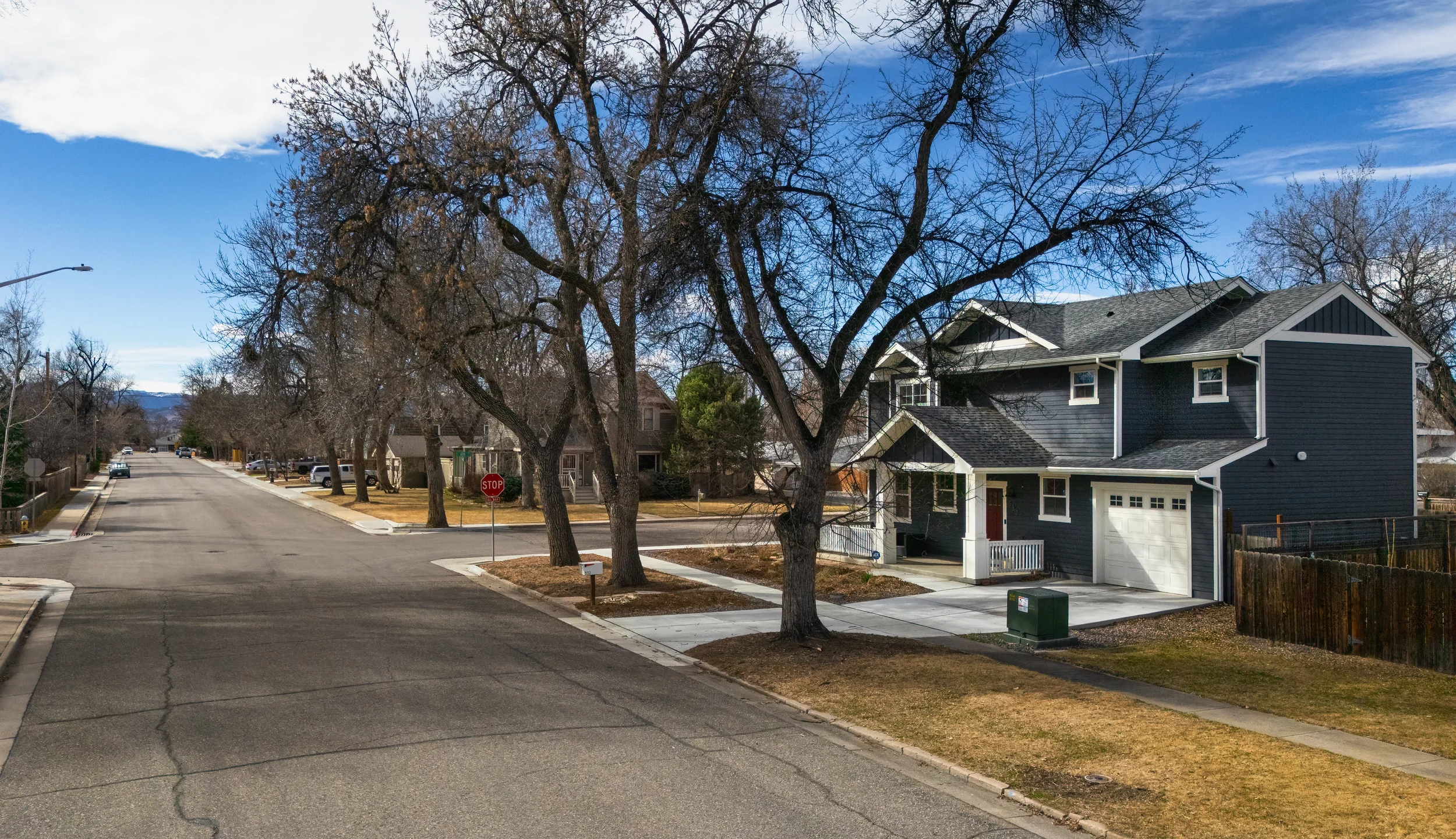 Aerial real estate photo taken in Berthoud, Colorado of a residential neighborhood street with a blue house on the right, leafless trees, a stop sign, and a clear sky.