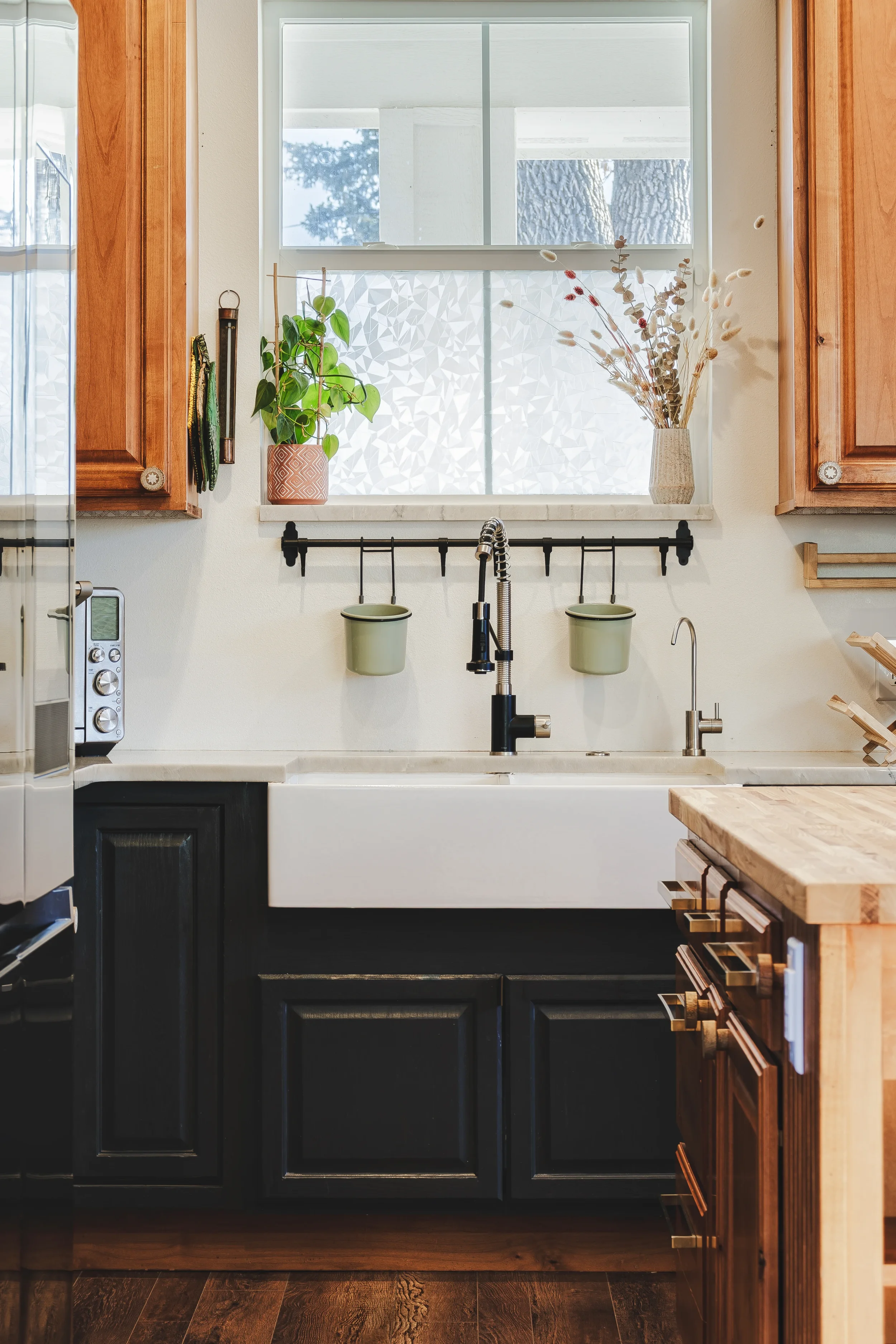 Close-up detail design photo of a white farmhouse sink, wooden cabinets, a window above the sink with decorative frosted glass, potted plants, and dried floral arrangements, black faucet, and a wooden countertop, taken in Berthoud, Colorado.