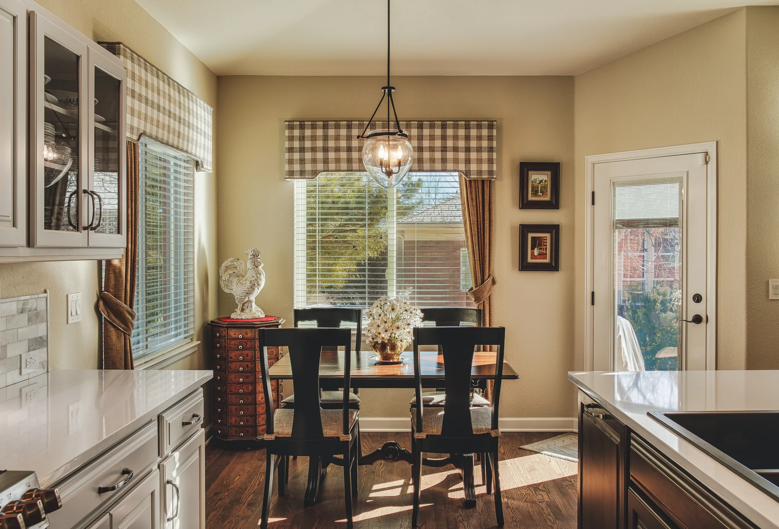 Architectural and Design photo of a cozy dining area in a kitchen with a dark wood table, a floral centerpiece, windows with valances and blinds, a door to a patio, and a rooster sculpture statue on a small red cabinet, taken in Longmont, Colorado.