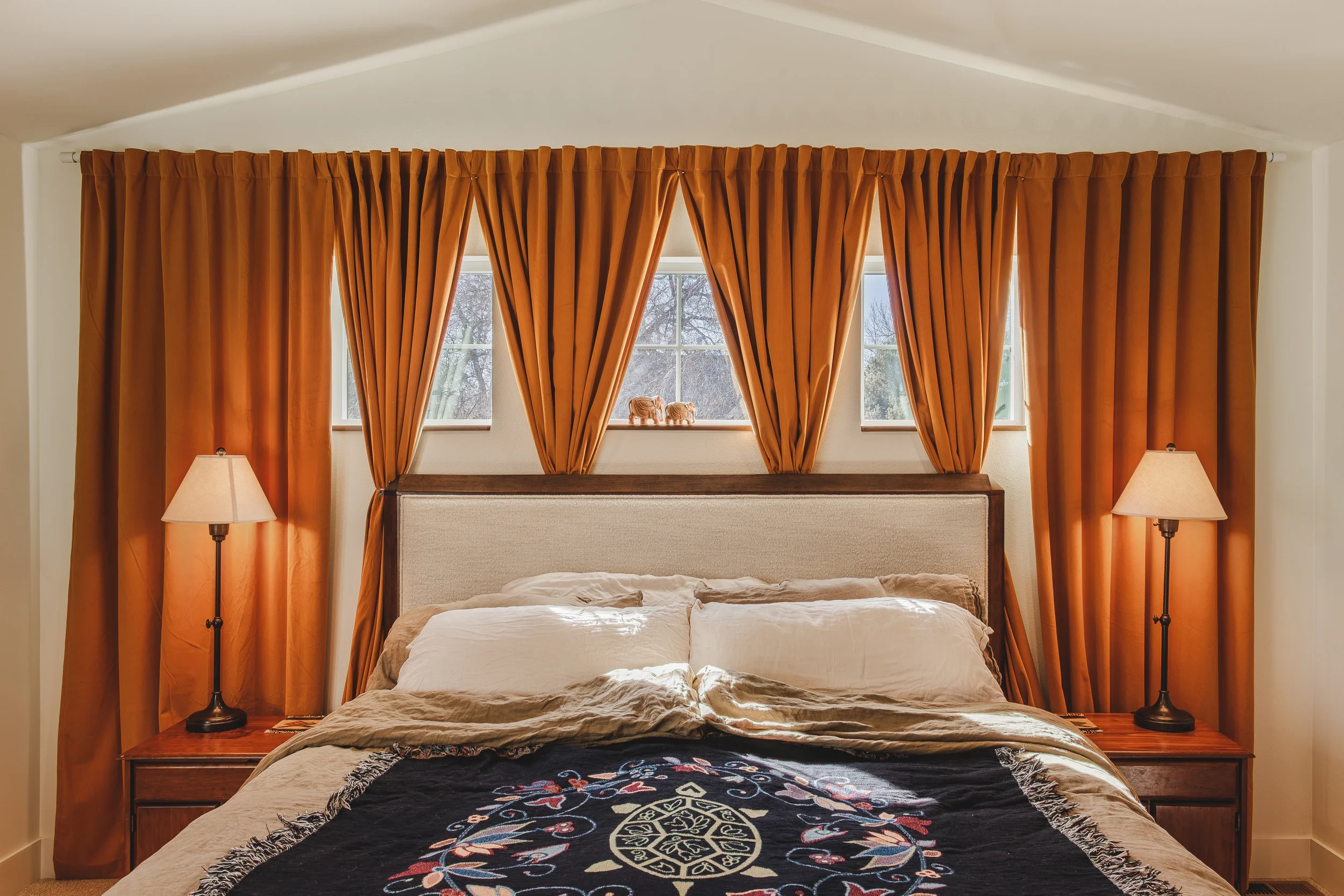 Interior Design photo of a master bedroom with large orange curtains hanging over a bed, slightly covering three windows with a winter scene outside, and sunbeams casting onto the bedsheets, shot in Berthoud, Colorado by Context Images. 