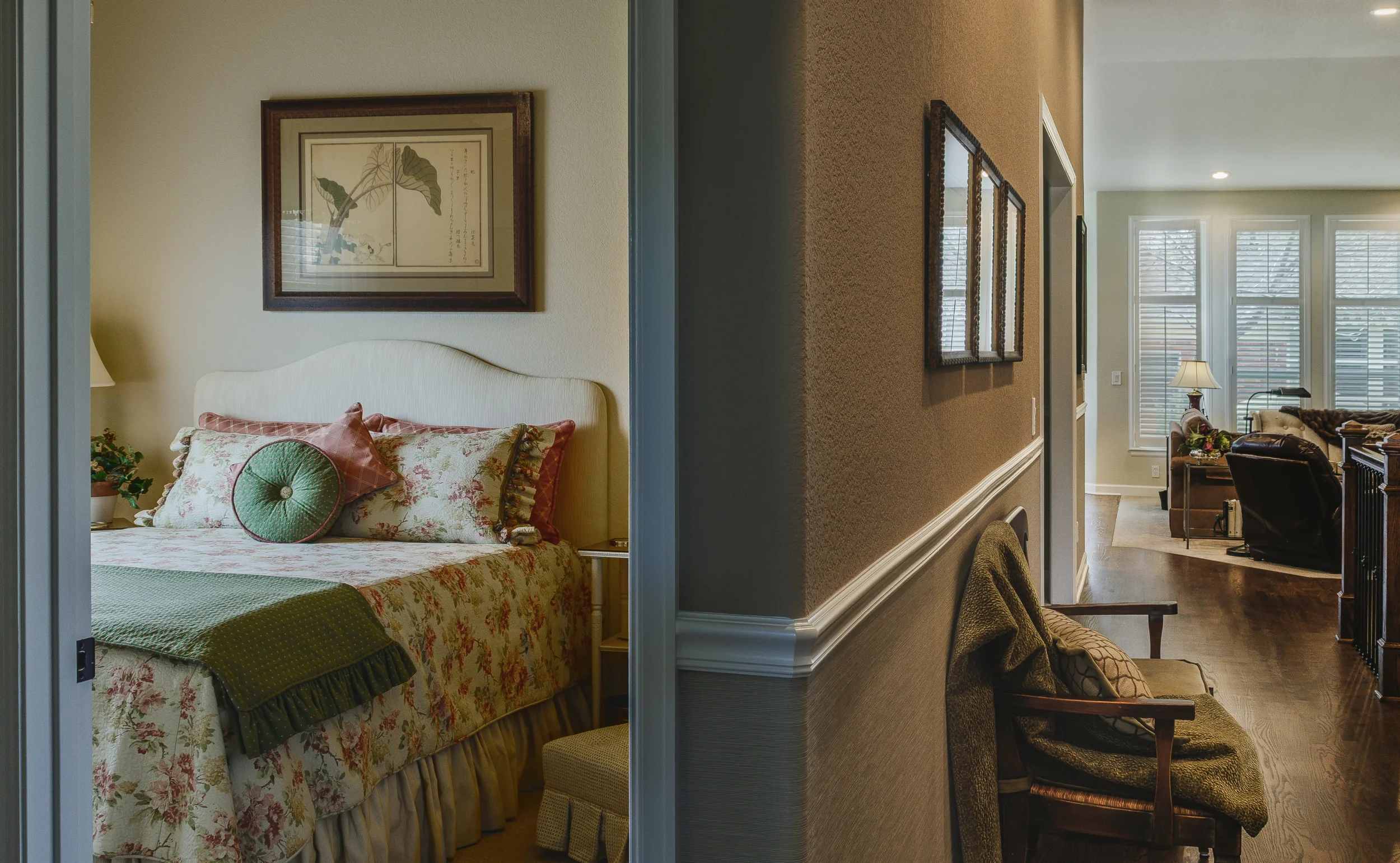 A publication level interior design and architecture photo of a bedroom on the left with floral bedding and framed artwork, and a hallway on the right leading into a living room with large windows. Taken in Longmont, Colorado.