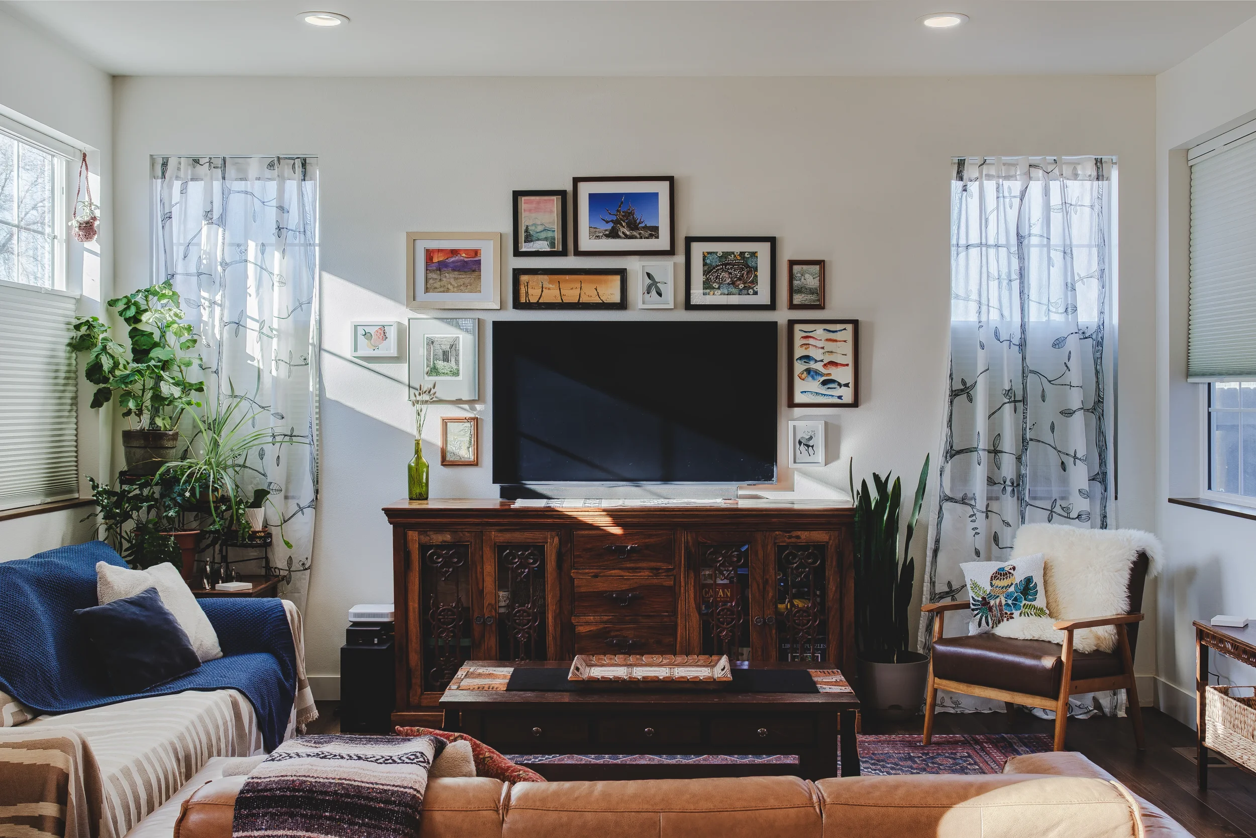 Eclectic living room with a Frame TV surrounded by a framed art gallery, leather sofa with cushions, armchair with a fur throw and decorative pillow, and potted plants near the window, shot by Context Images in Berthoud, Colorado.