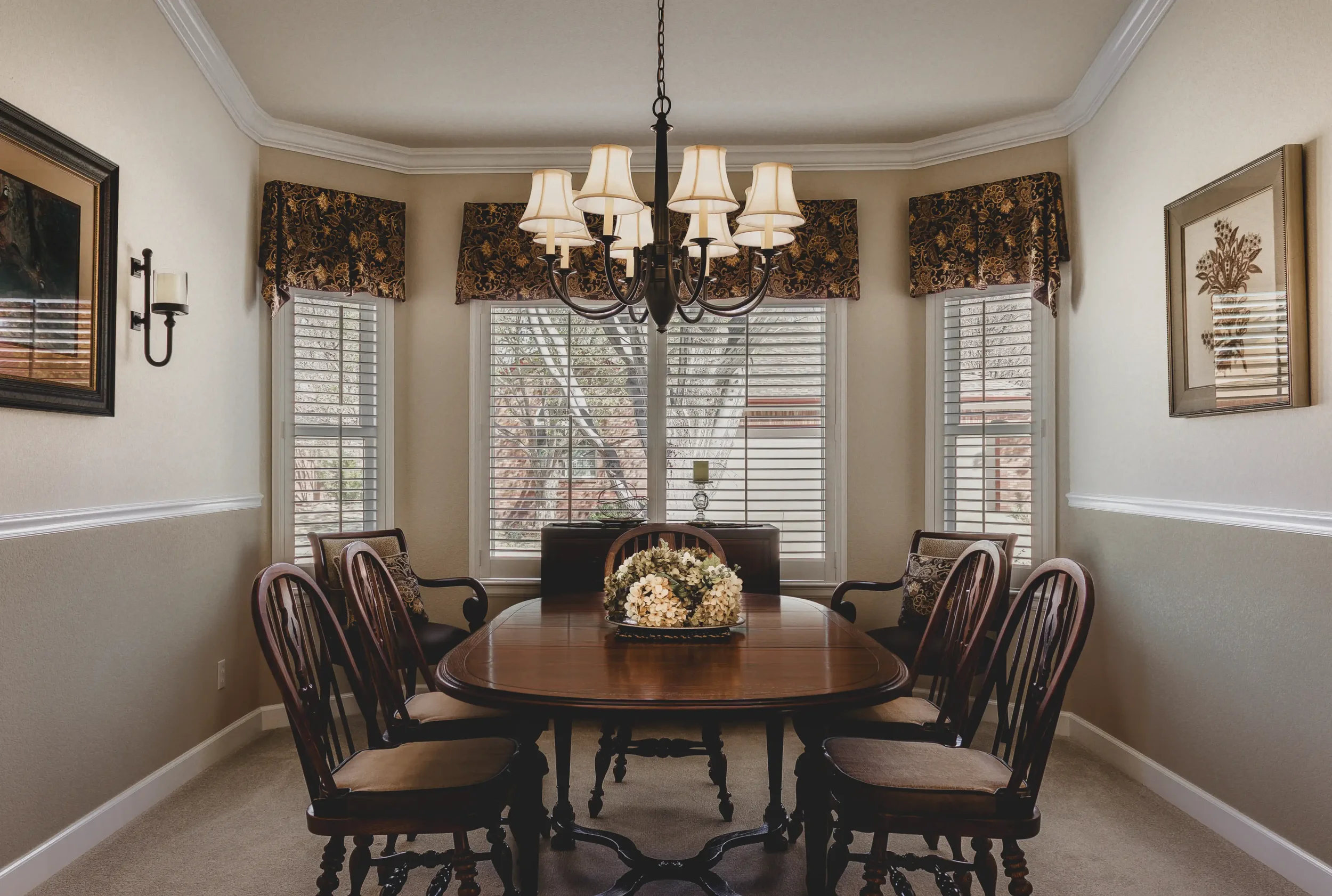 A cinematic interior shot of a dining room with an oval wooden table, six chairs with cushioned seats, a chandelier hanging from the ceiling, and windows with floral valances and white blinds, shot in Longmont, Colorado.