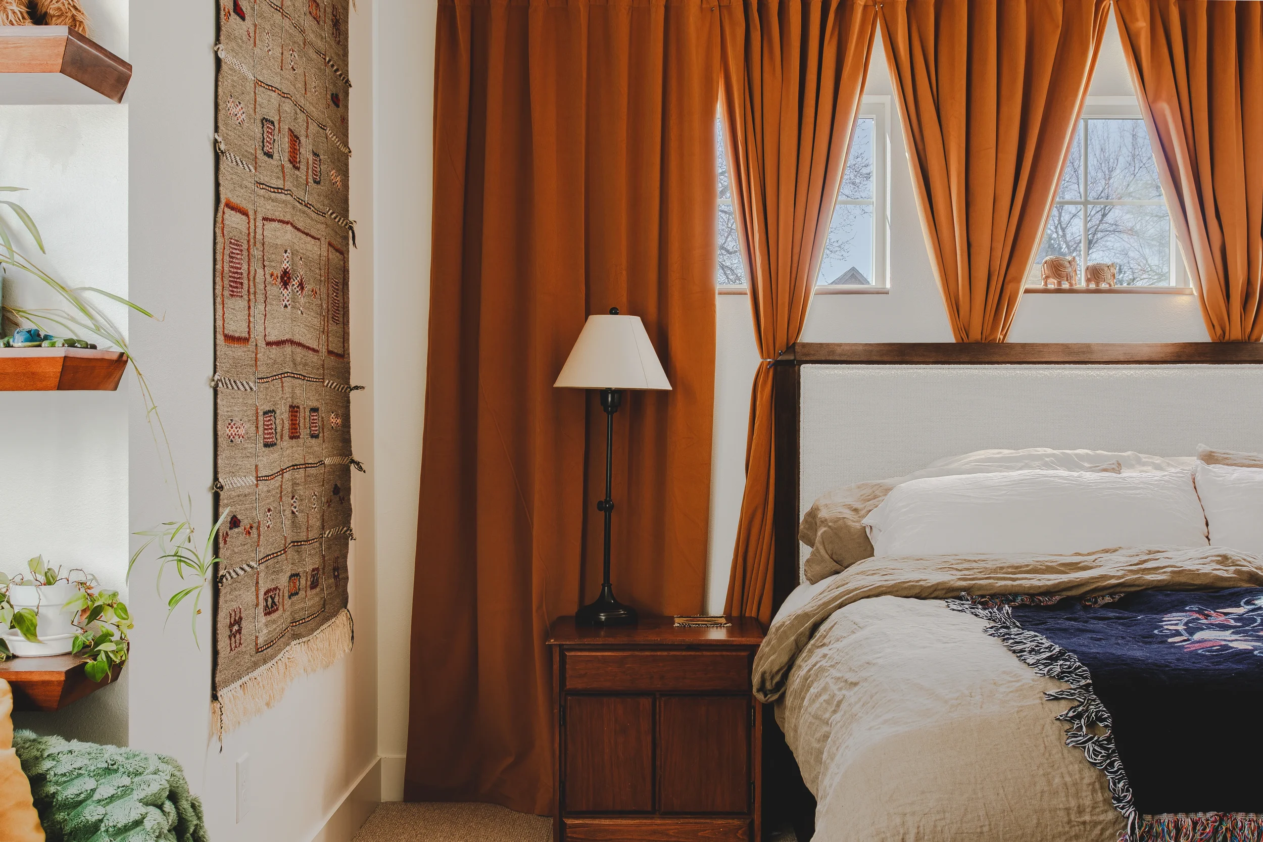 A cozy bedroom corner with a bed, orange curtains, a wooden nightstand, and a lamp near a window, photographed in Berthoud, Colorado.