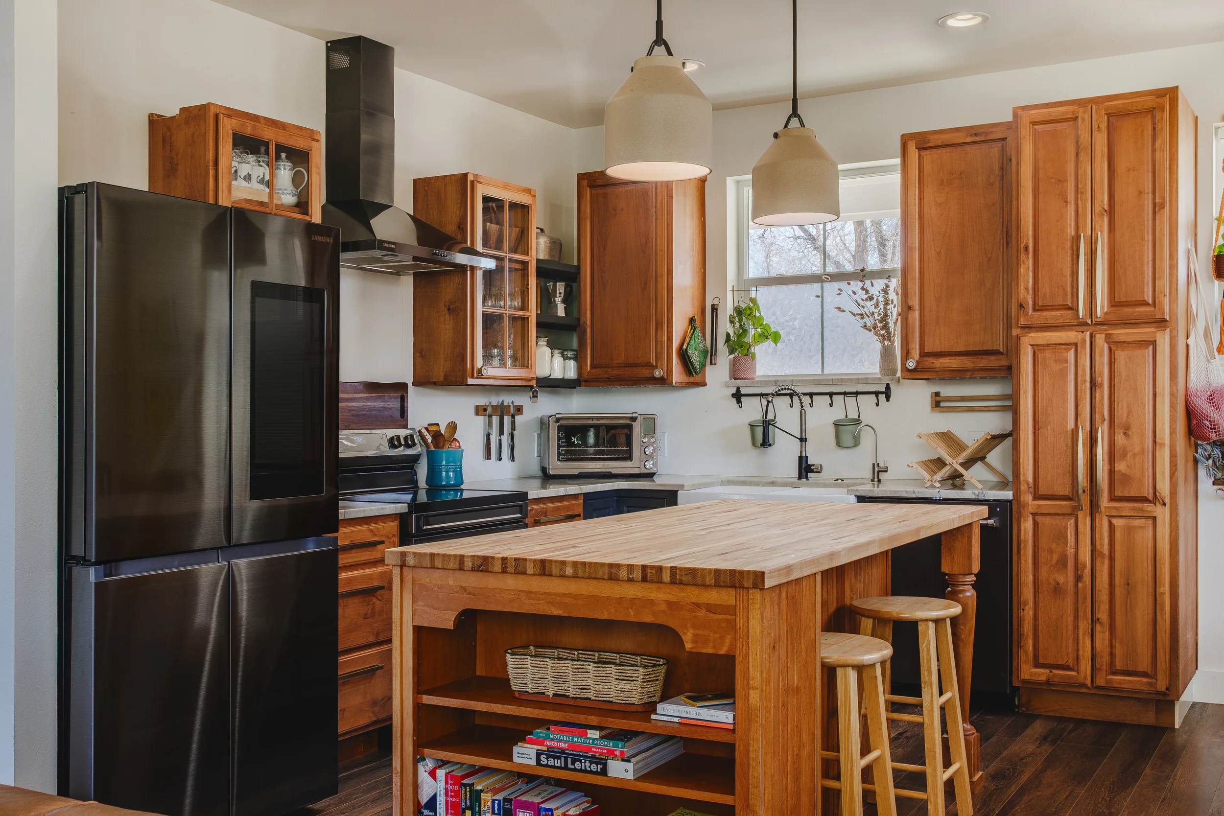 Architectural & design photo of kitchen with wooden cabinets, a stainless steel refrigerator, a black stove, a small countertop oven, a butcher block island, and beige pendant lights, taken in Berthoud, Colorado by Context Images.