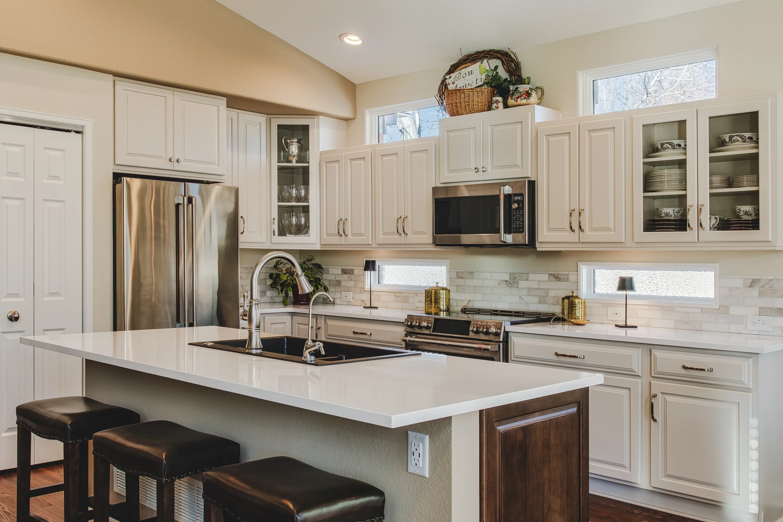 Architectural and Design photo of a modern kitchen with white cabinets, stainless steel appliances, an island with black sink and chrome faucet, and decorative items on the countertops. Windows let in natural light. Taken in Longmont, Colorado.