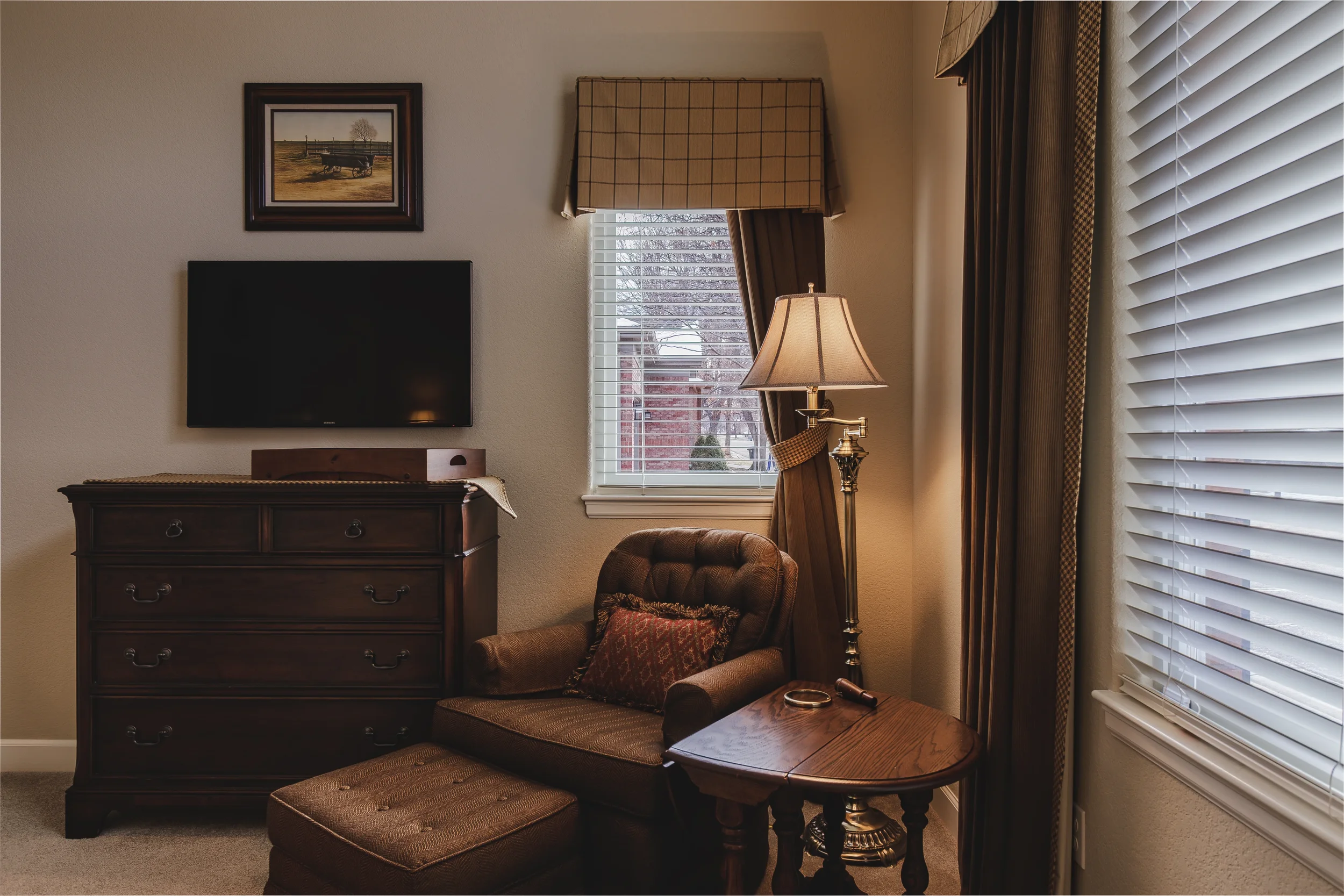 An interior design photograph of a cozy bedroom corner with a wooden dresser, a wall-mounted flat-screen TV, a brown upholstered armchair with a patterned cushion, a small wooden side table with a wristwatch and eyeglasses, a floor lamp with a beige 