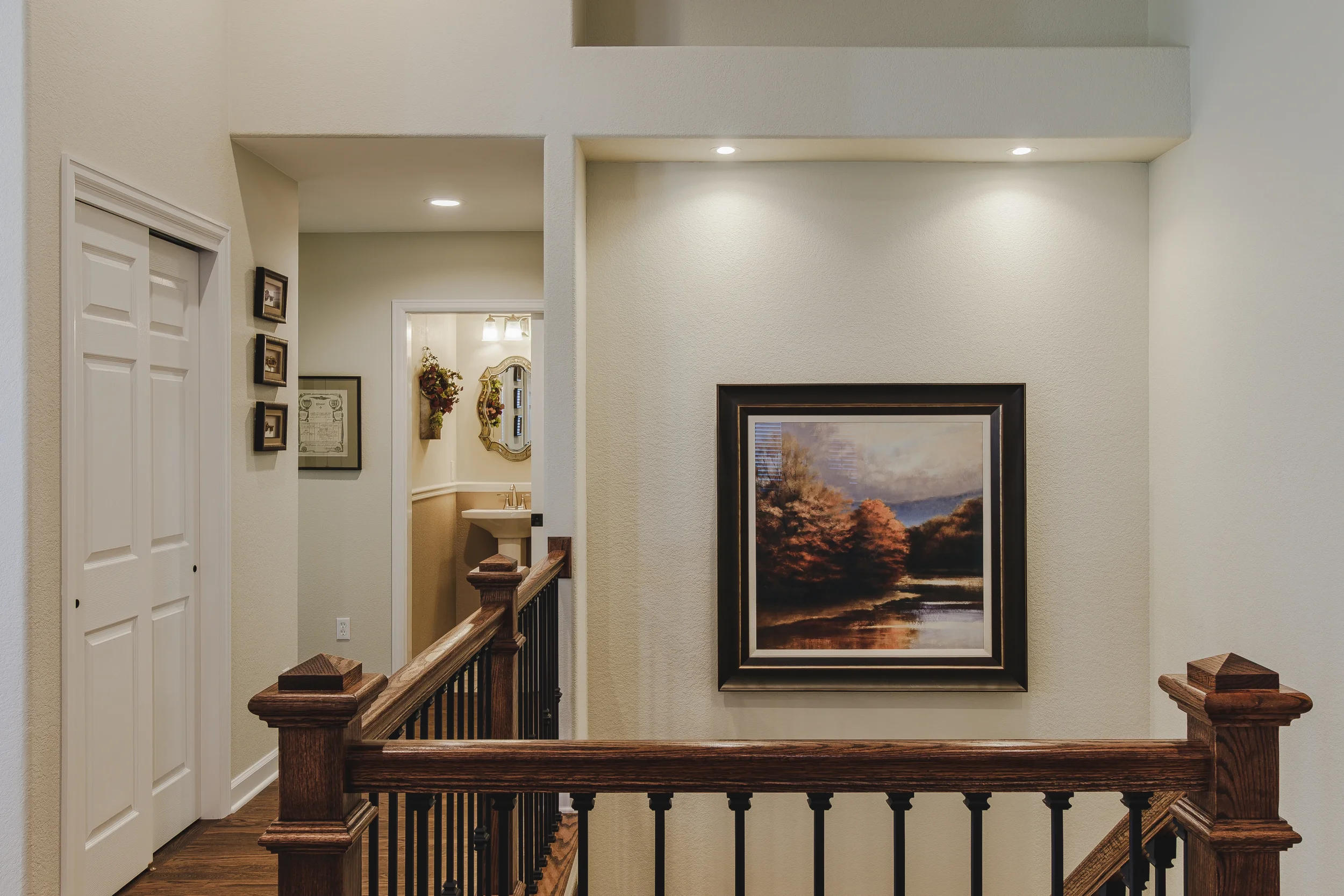 Interior view of a first-floor hallway with a wooden railing, a large framed landscape painting, and a bathroom with a sink and mirror in the background, taken in Longmont, Colorado by Context Images. 