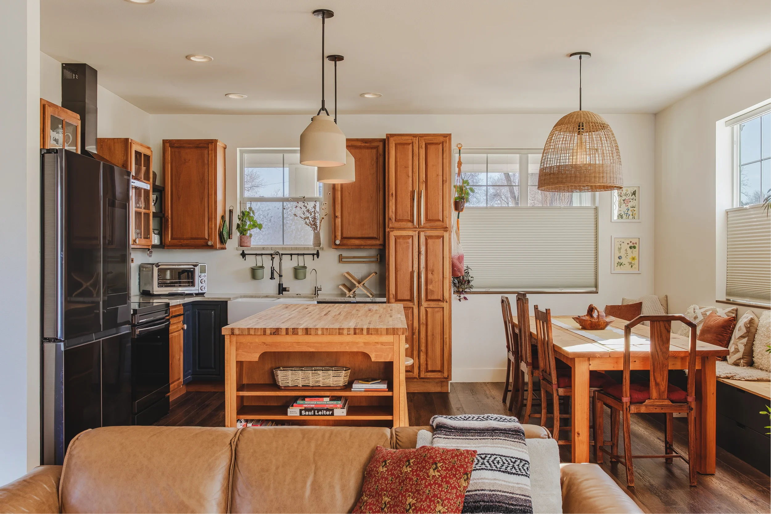 A warm open-concept kitchen and dining area with wooden cabinets, black appliances, a butcher block island, and a dining table with six chairs, plus hanging pendant lights and window blinds, taken in Berthoud, Colorado by Context Images.