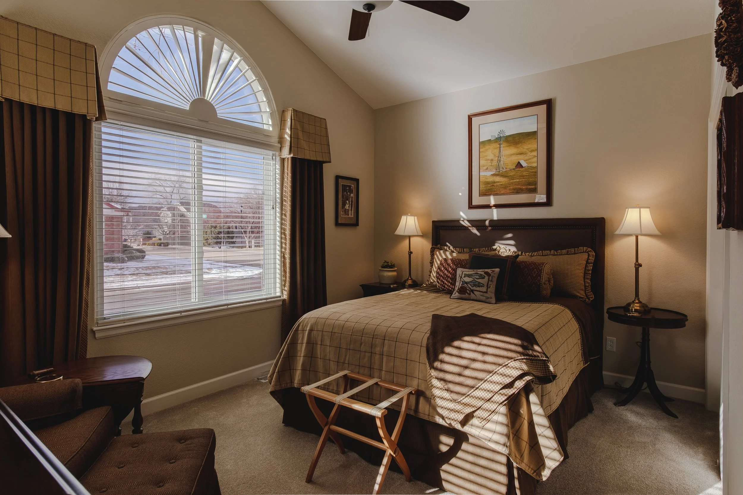 A cozy bedroom with a large window, two bedside lamps, a framed landscape painting above the bed, and a luggage rack at the foot of the bed. Taken in Longmont, Colorado by Context Images. 