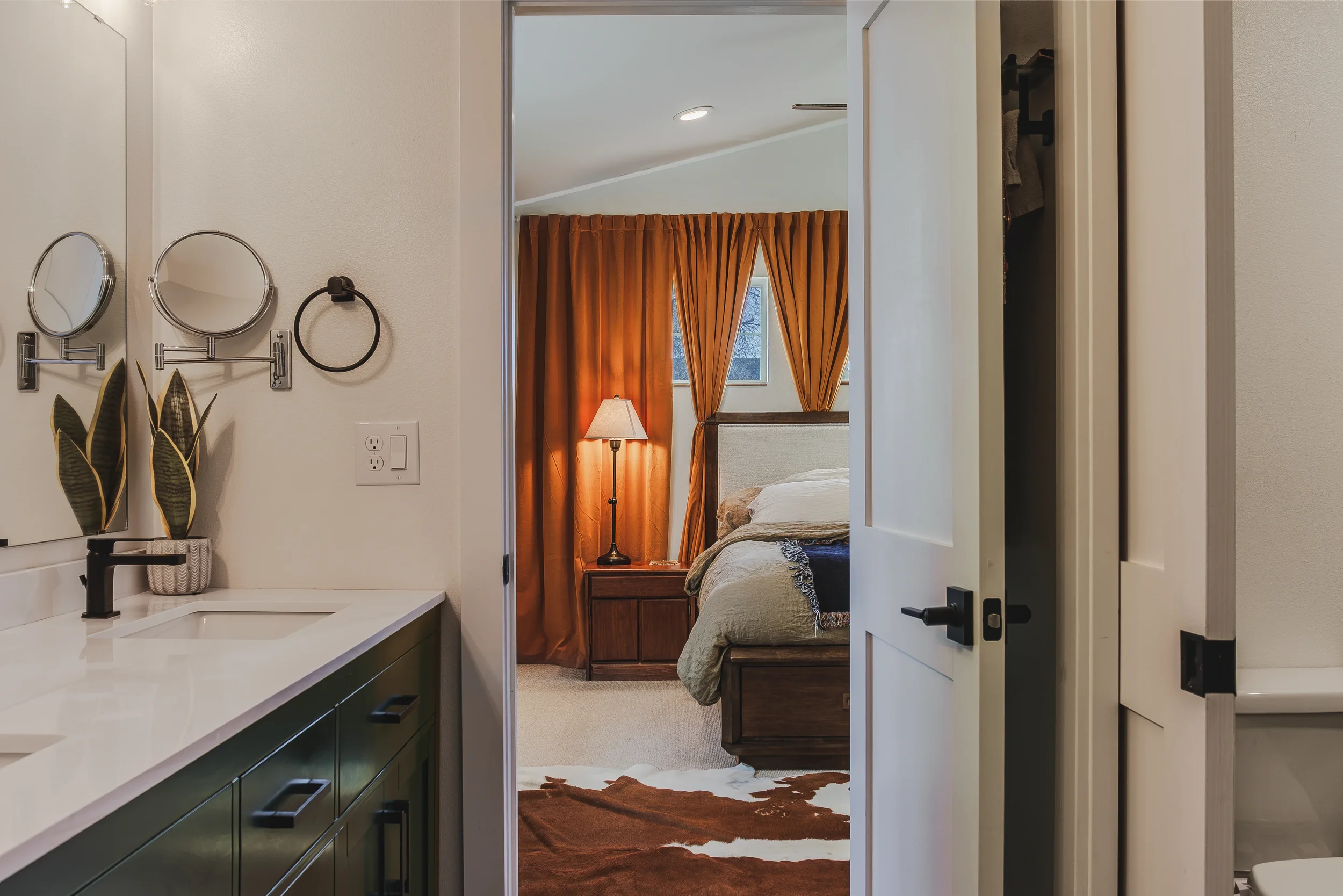 First-person view from a bathroom looking into a bedroom with orange curtains, a bed with a beige bedspread, and a wooden nightstand with a lamp, taken in Berthoud, Colorado. 