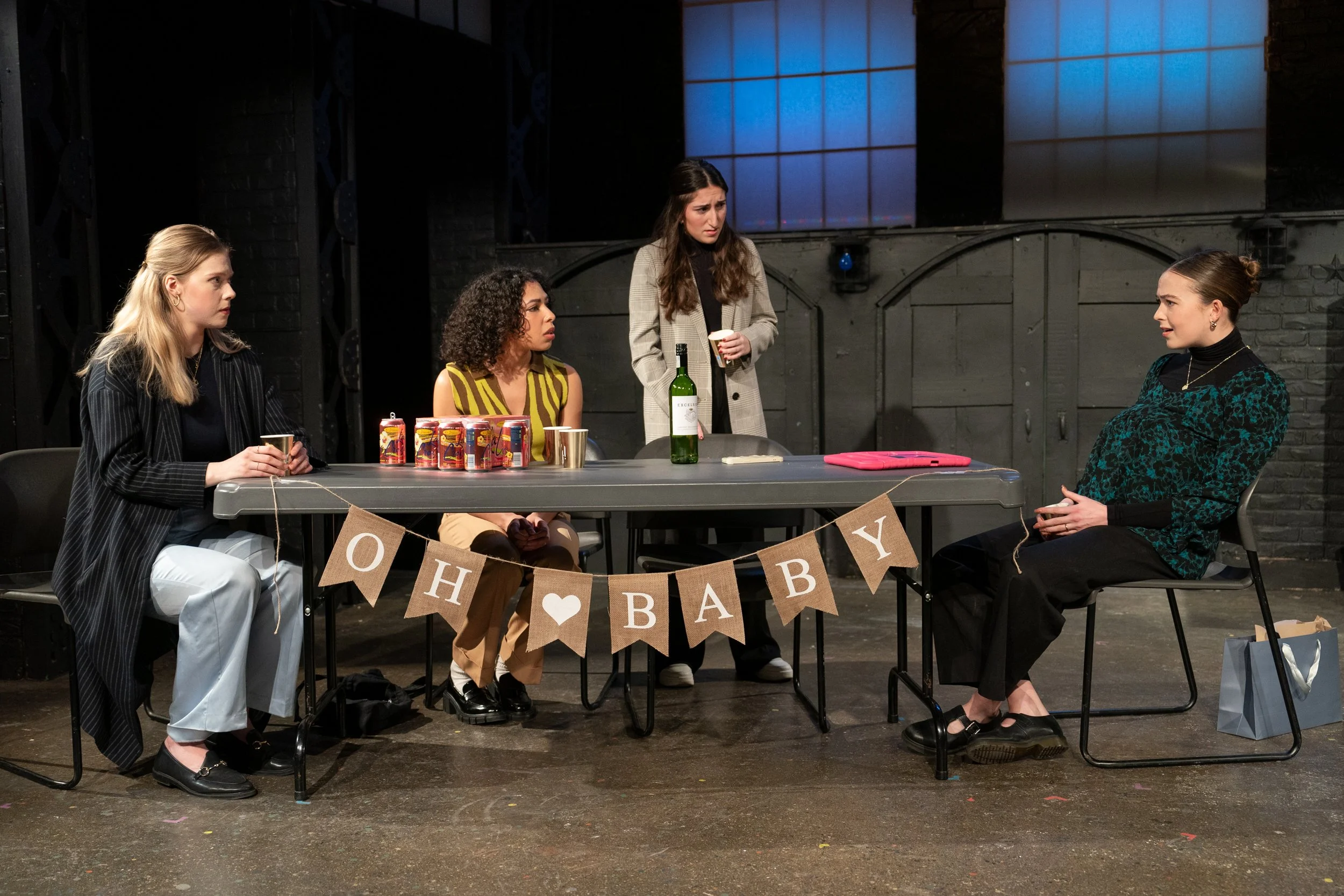 Four women sitting at a table with drinks and snacks, one standing, in a room with dark brick walls and aHappy Baby banner hanging on the front of the table.