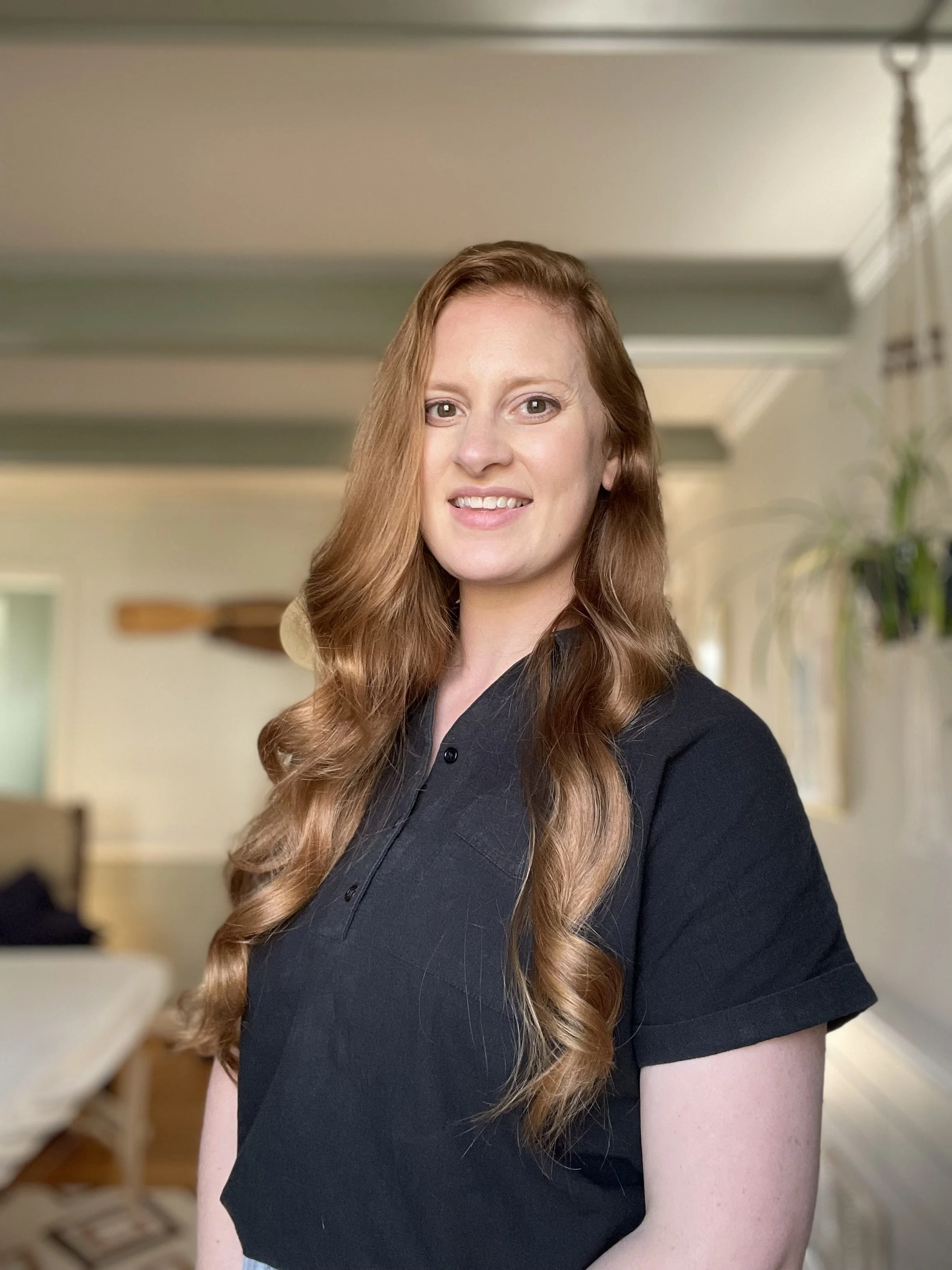 A young woman with long, wavy red hair, smiling, wearing a black shirt, standing in a well-lit room with green walls and a plant in the background.