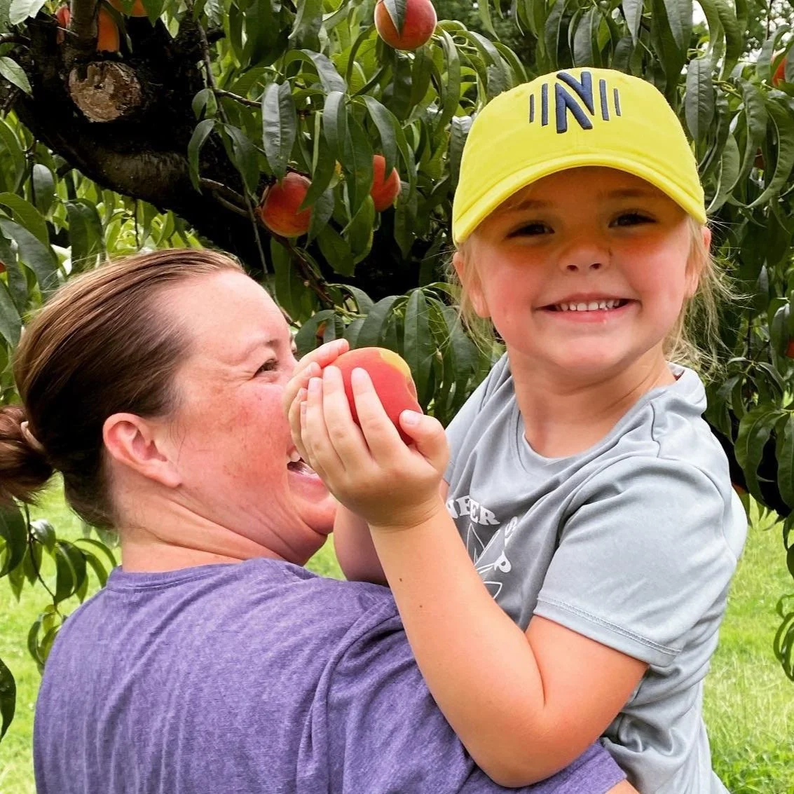 A woman holding a young girl in an apple orchard, with the girl holding a peach and wearing a yellow cap, both smiling.