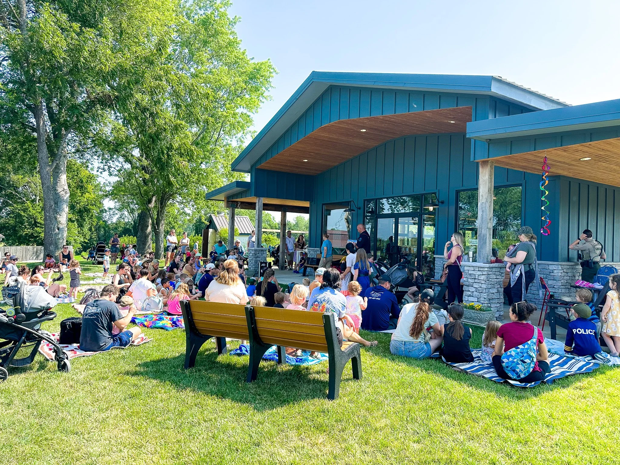 A large outdoor gathering with many people sitting on the grass and benches, attending a concert or event on a stage outside a blue building with wood accents, surrounded by green trees on a sunny day.