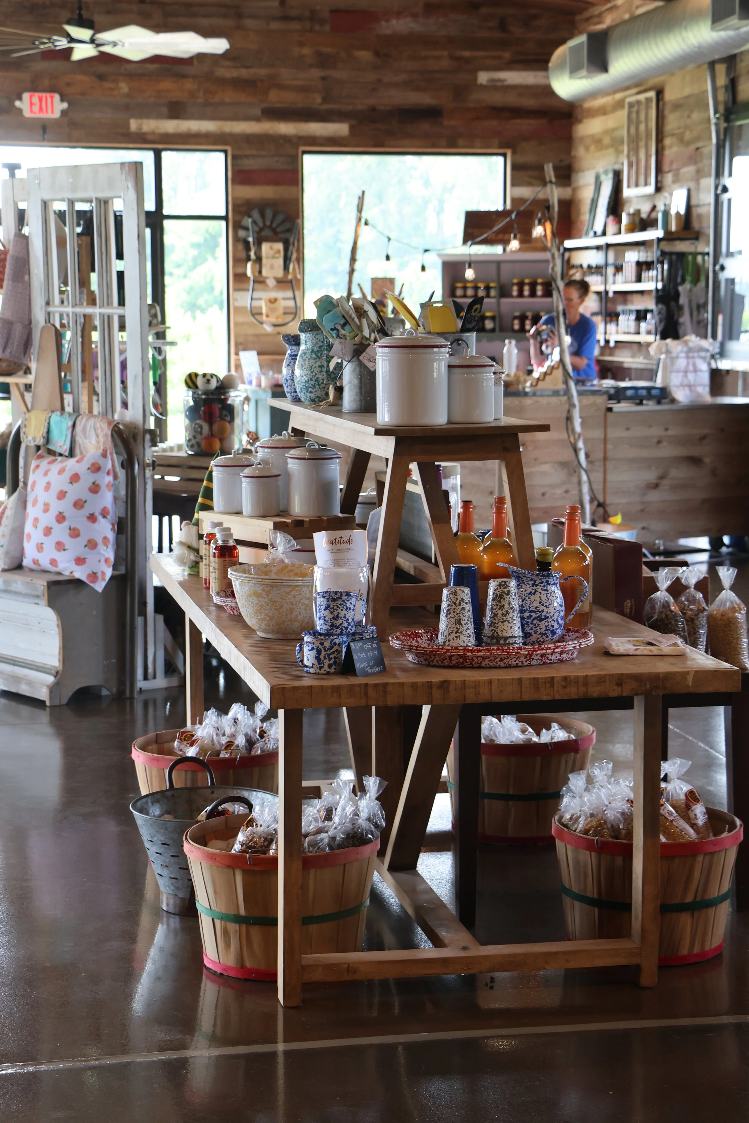 Interior of a small retail shop with wooden walls and shelves, displaying various products including ceramics, bottles, and packaged goods. A person is seen working behind the counter, with large windows letting in natural light.