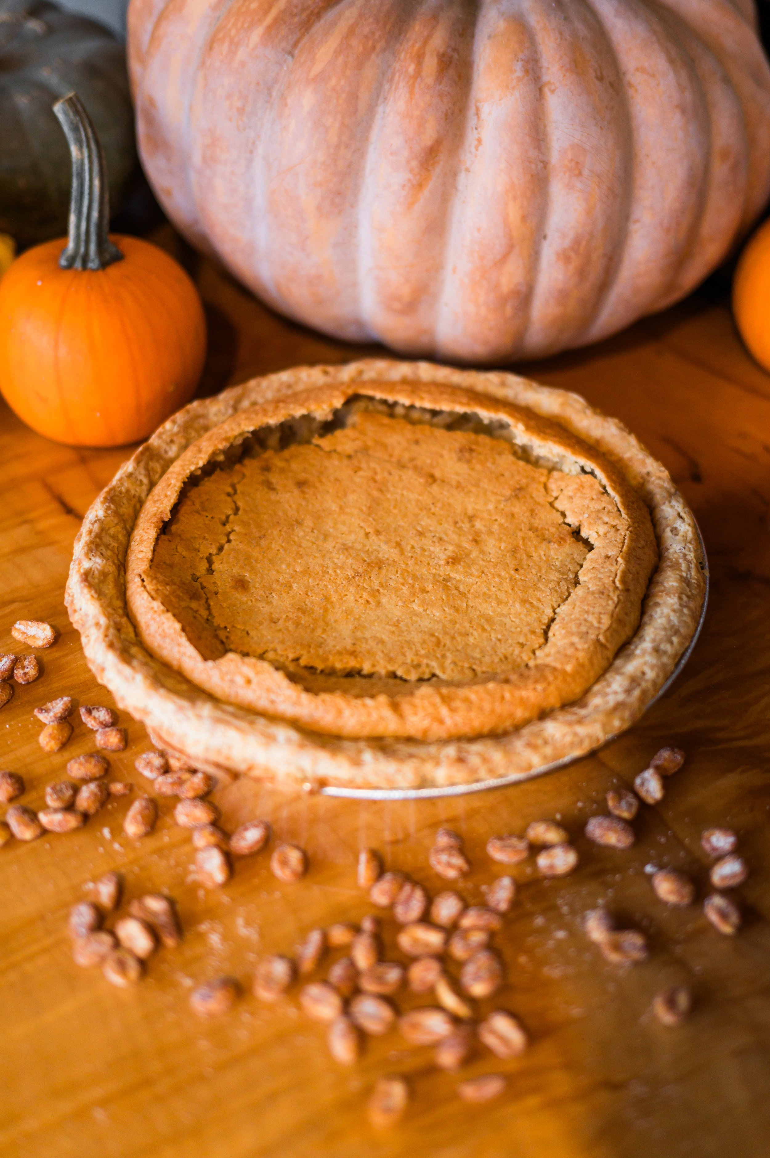 A pumpkin pie with a golden crust, surrounded by small pumpkins and a large pumpkin, on a wooden surface with scattered grains.