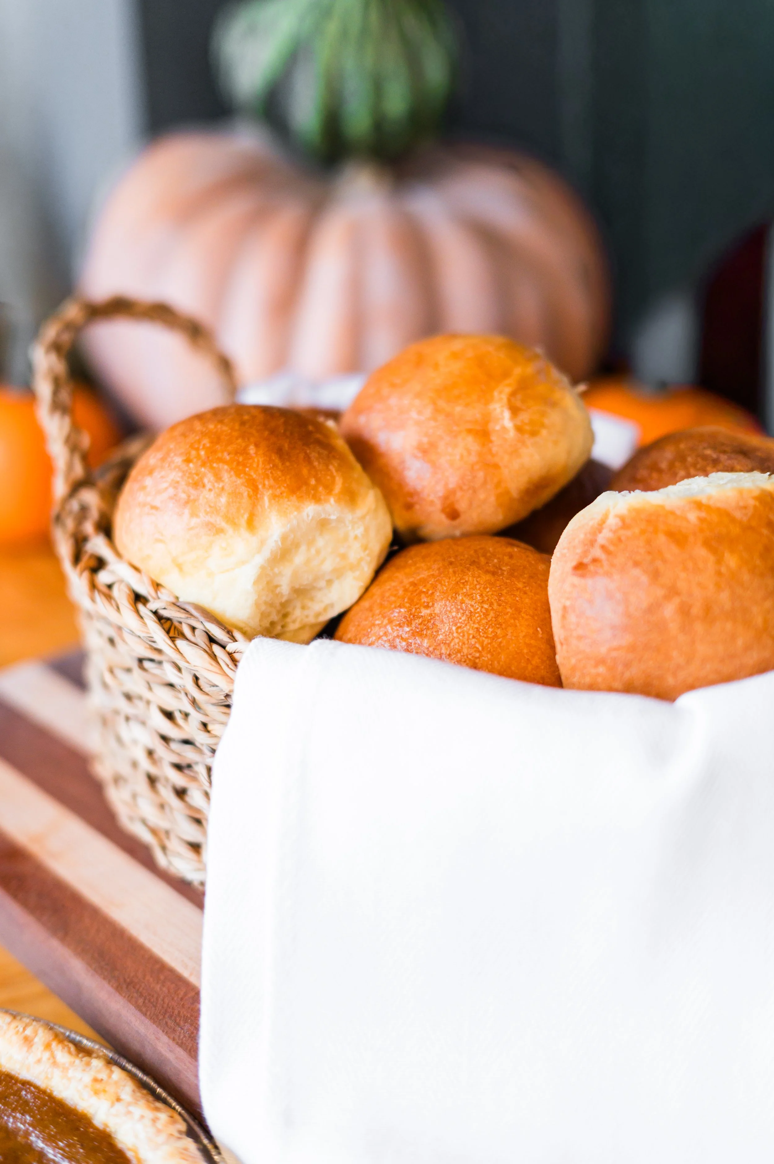 A basket of freshly baked bread rolls on a wooden surface with a white cloth lining, with decorative pumpkins in the blurred background.