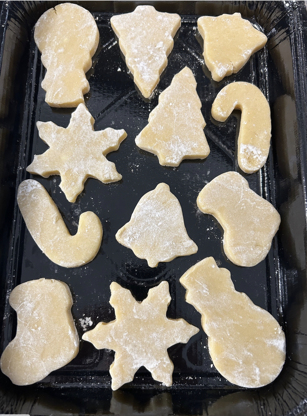 Unbaked sugar cookies shaped like Christmas trees, snowflakes, candy canes, and stockings on a baking sheet with a dusting of flour.