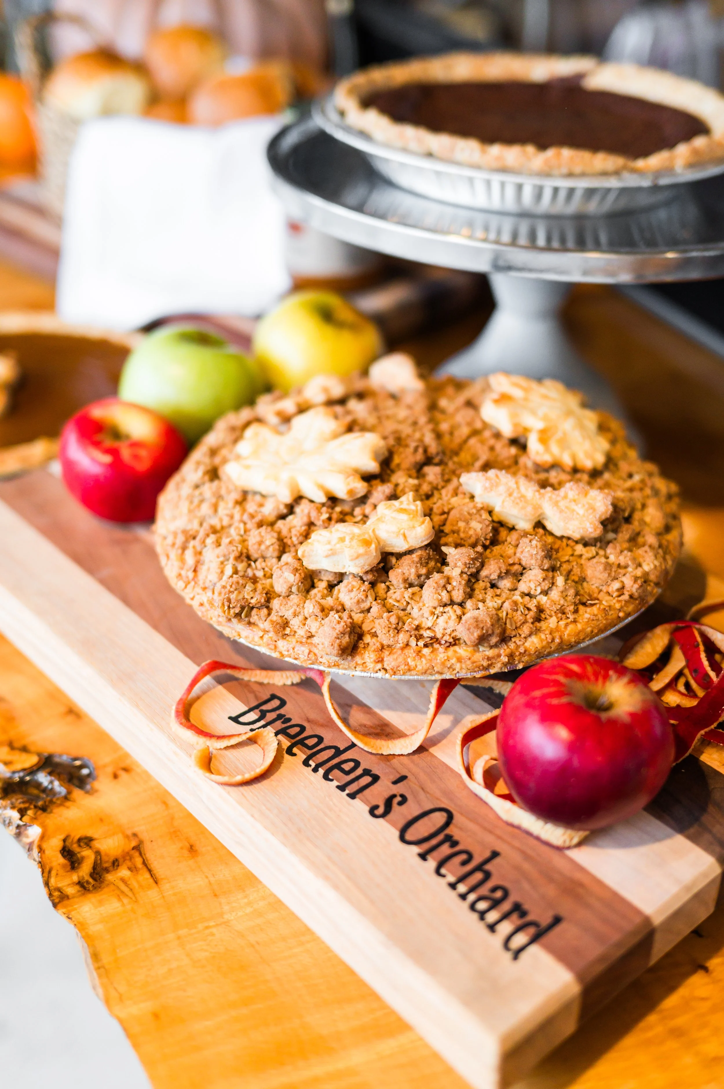 Apple crumb pie on a wooden board with red and green apples and a pumpkin pie in the background.