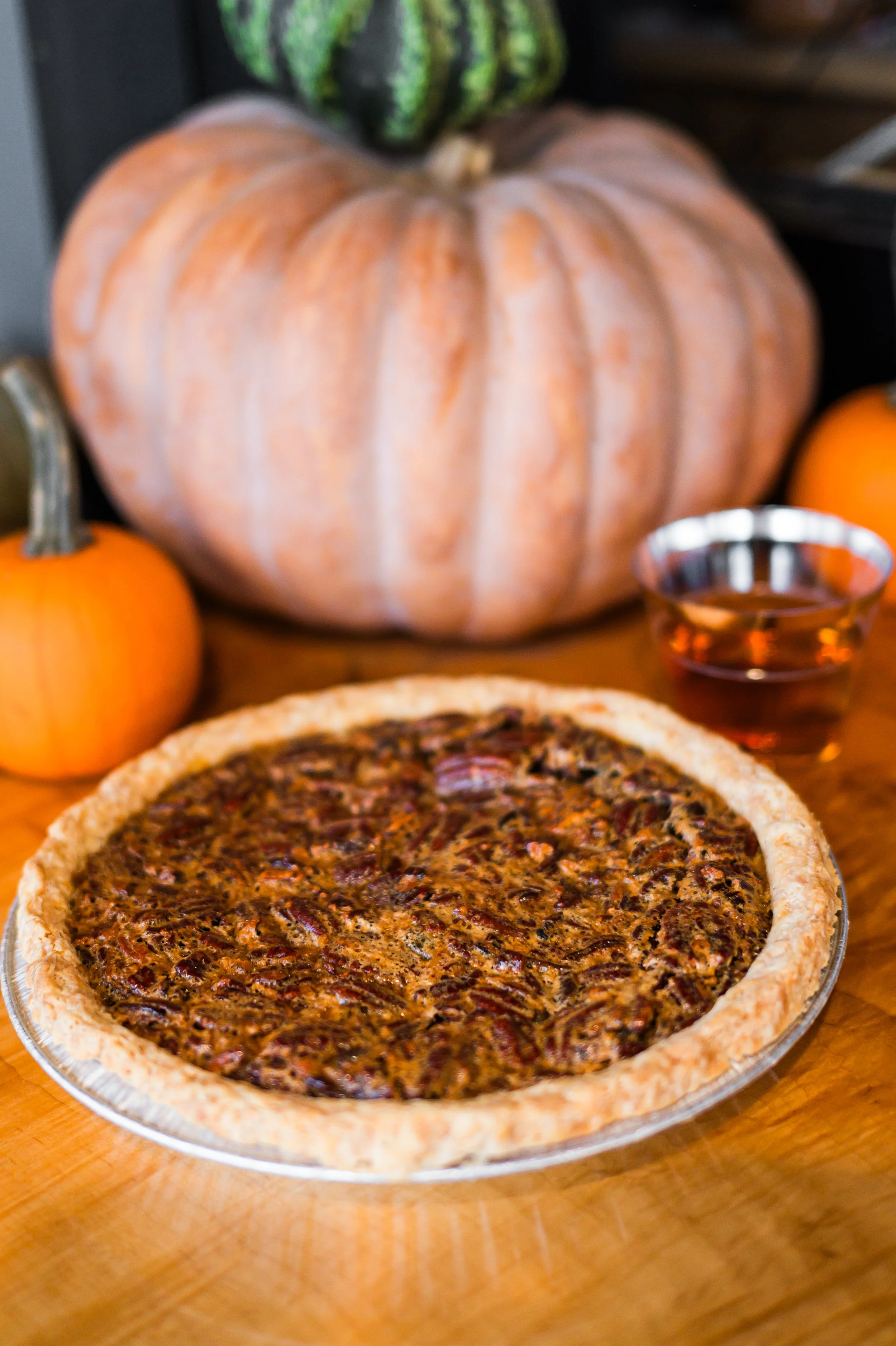 A pumpkin pie on a wooden table with a large pumpkin, small pumpkin, and a glass of tea in the background.