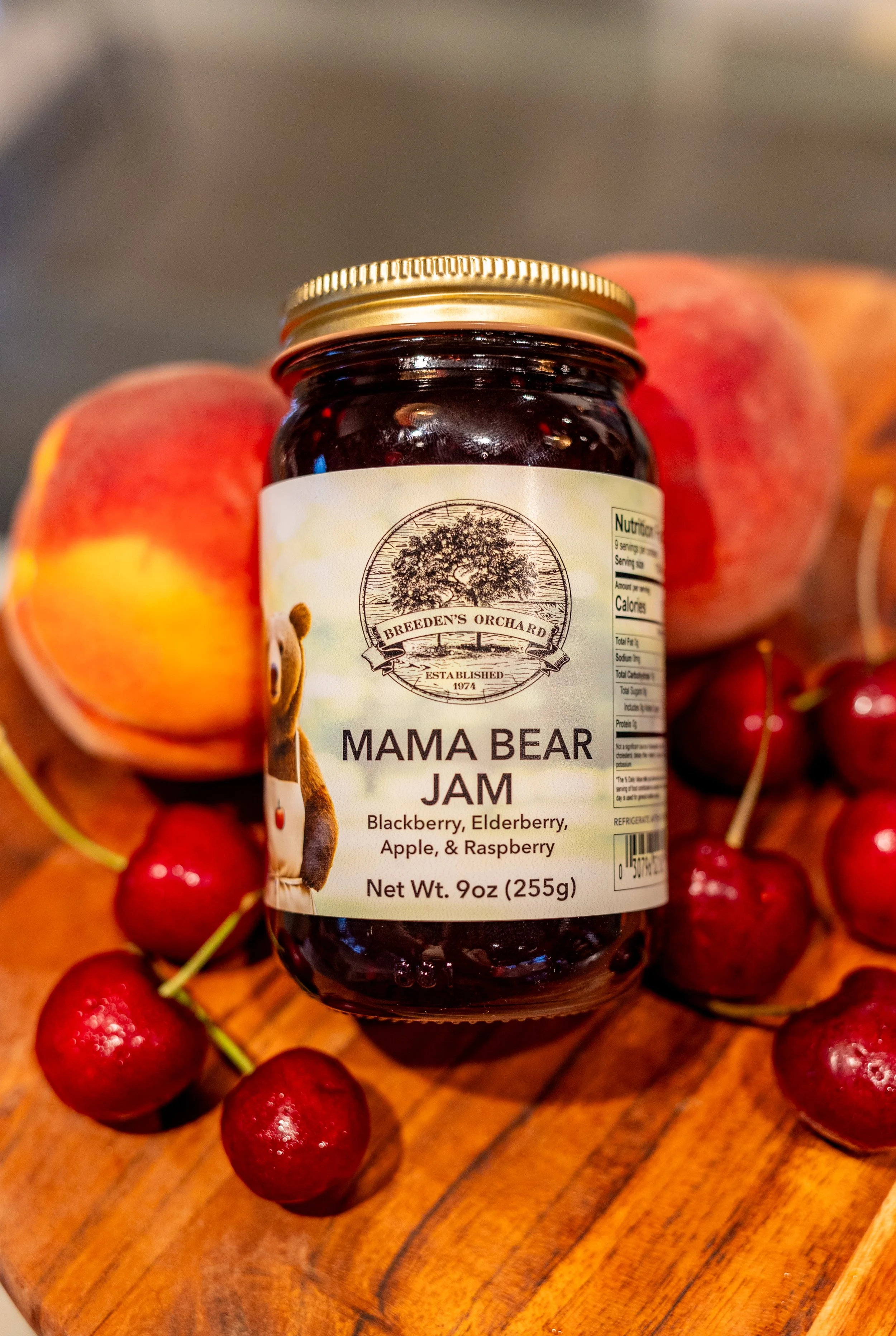 A jar of Breedens Orchard Mama Bear Jam, flavored with blackberry, elderberry, apple, and raspberry, surrounded by fresh peaches and cherries on a wooden surface.