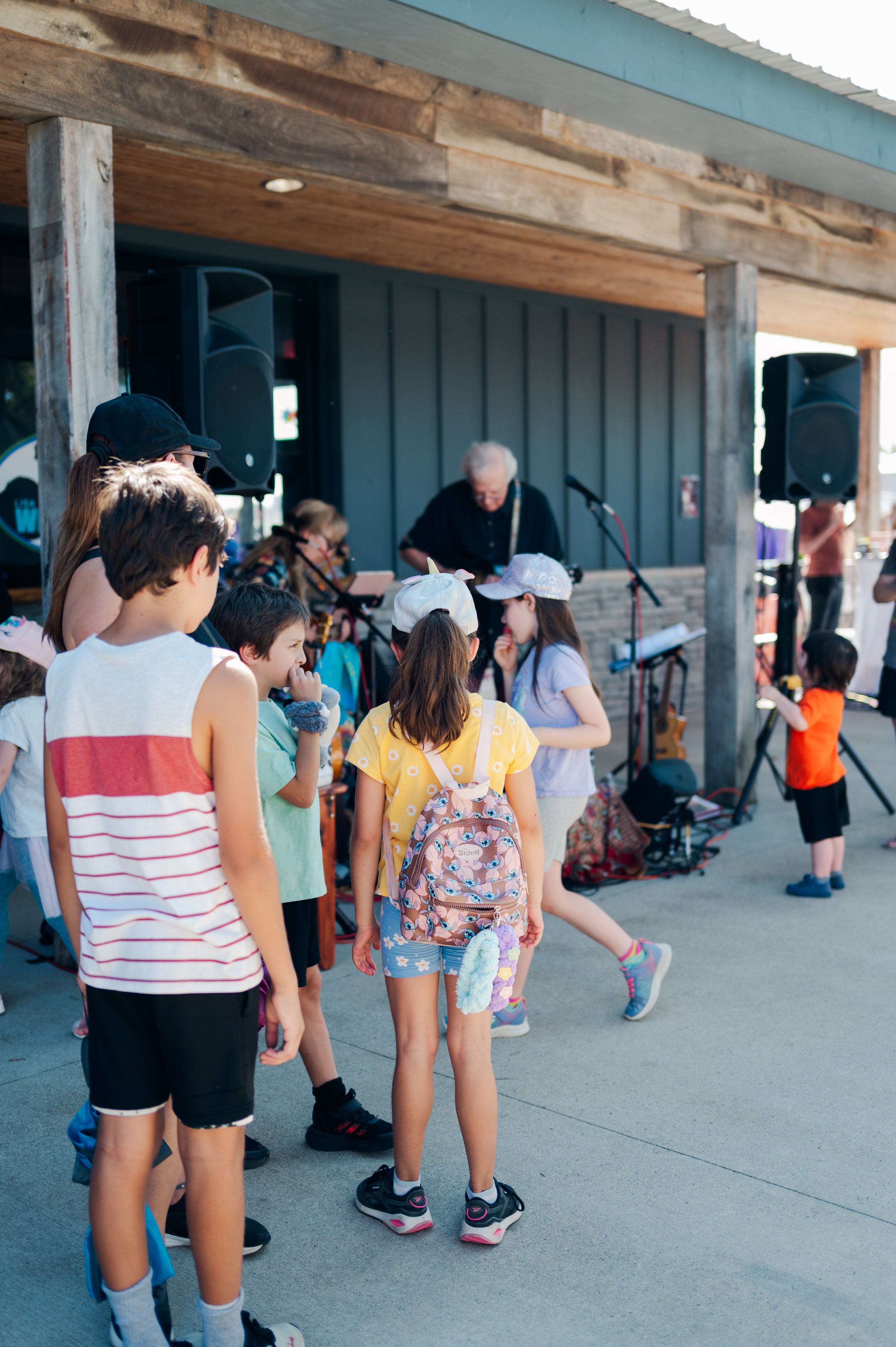 Children gathered outdoors near a stage, watching a live music performance by a man with long hair and glasses. The children are wearing casual summer clothes and backpacks.