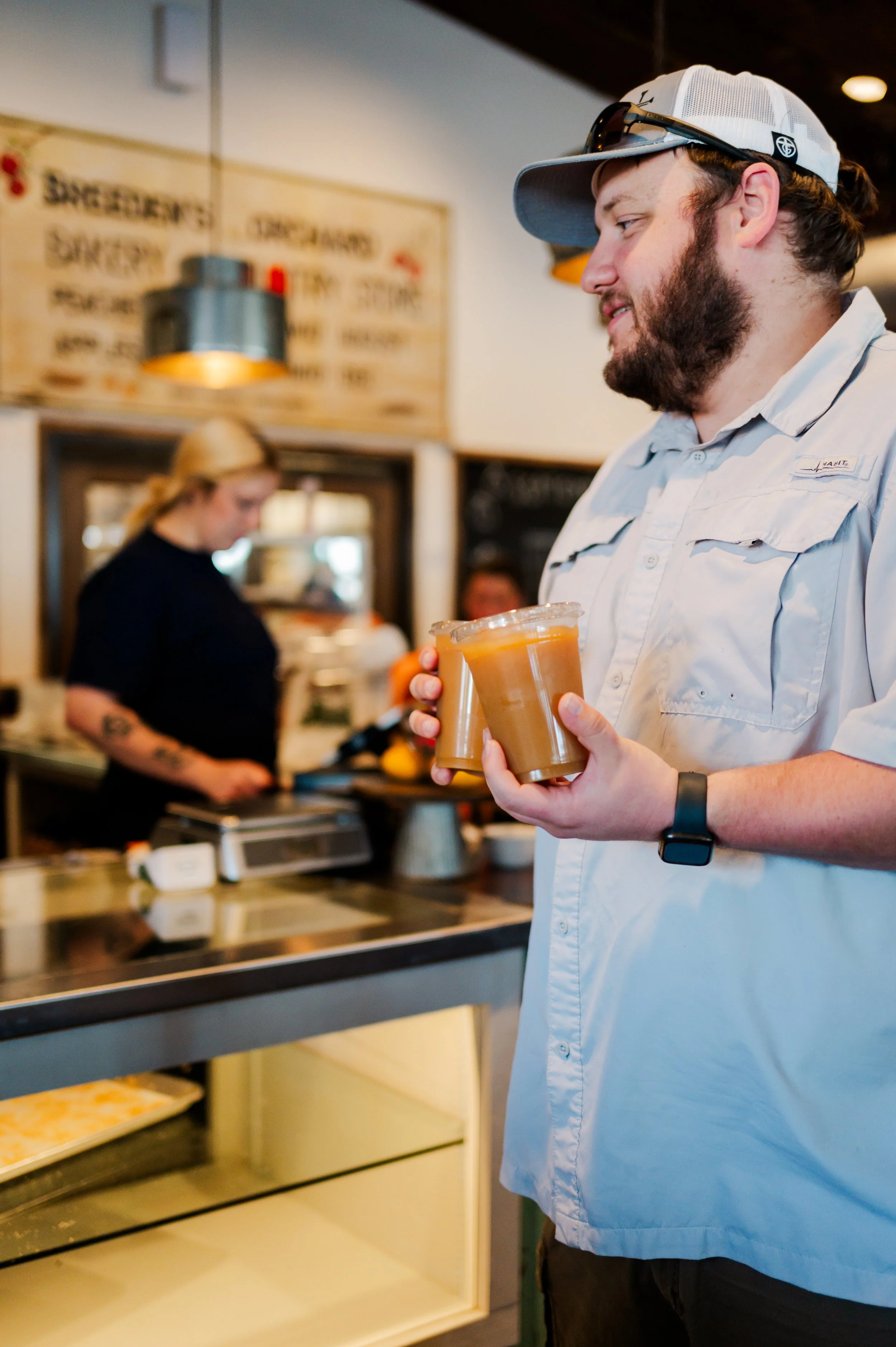 A man in a white shirt, baseball cap, and sunglasses holds two cups of coffee in a café, with a woman behind the counter preparing orders in the background.