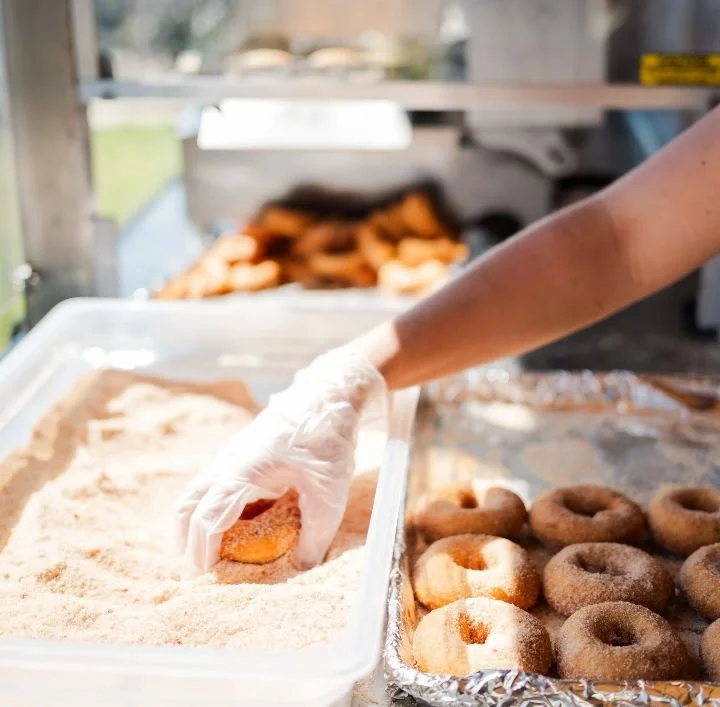 A baker wearing a glove is coating donut holes with powdered sugar in a white container, with more donuts on a tray covered with foil nearby.