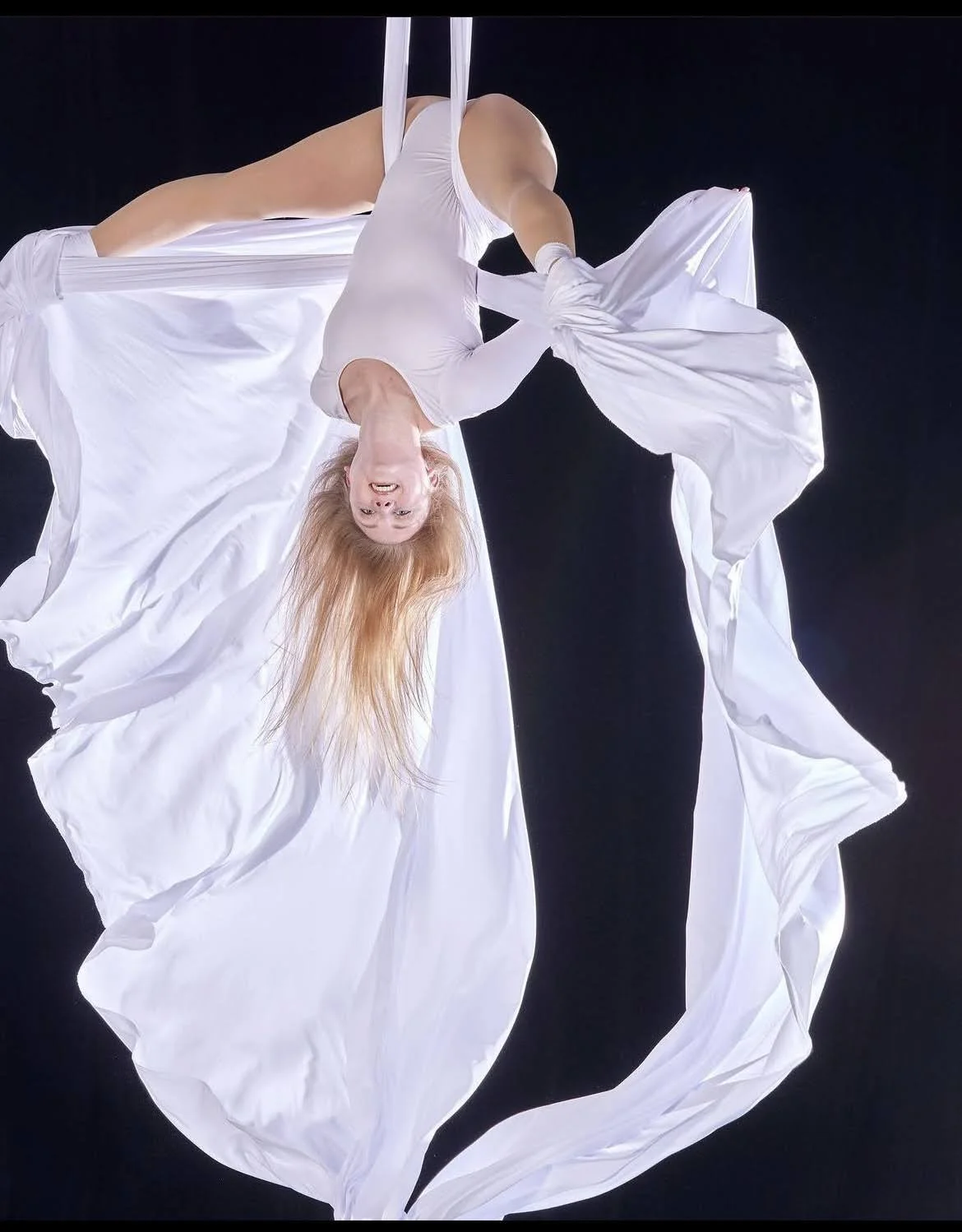 A woman with long red hair is hanging upside down against a black background, wearing a white outfit with flowing fabric, and smiling at the camera.