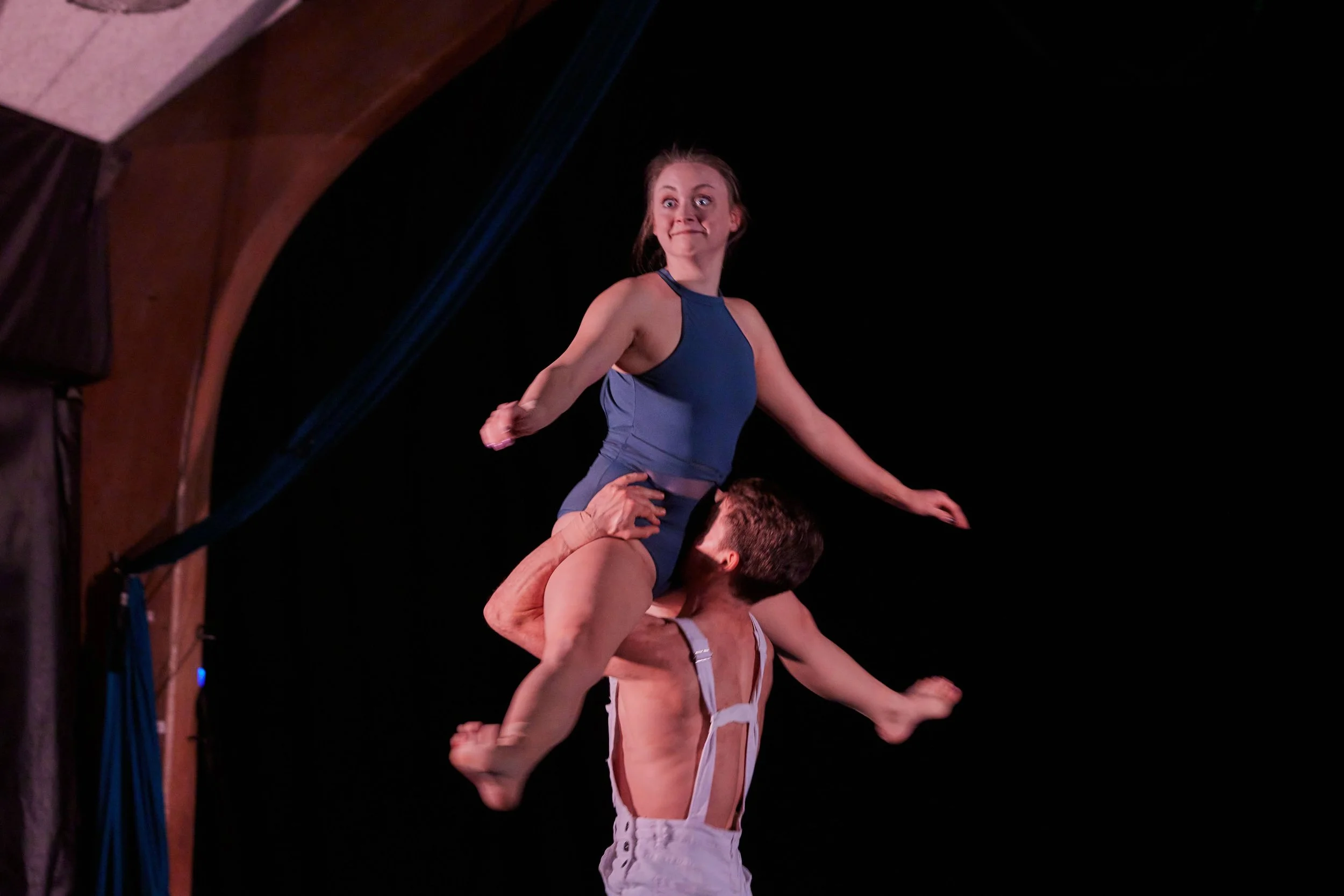 A male acrobat wearing white suspenders supports a female acrobat dressed in blue, who is sitting on his shoulders, during a performance against a dark background.